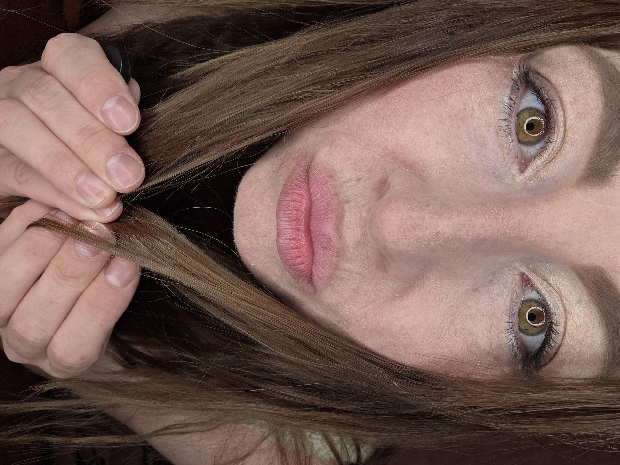 This image depicts a close-up, eye-level view of an adult female located in Chaikovsky, Russia. Her face, characterized by light skin tone with visible texture and minor lines, occupies the central portion of the frame. She has hazel eyes, which are looking directly at the viewer, with visible eyeliner and a shimmering, light-toned eyeshadow. Her lips are pursed into a distinct pout. Long, straight or slightly wavy light brown hair frames her face, with strands held forward by her hands below her chin. Her hands, with natural, unpolished fingernails showing lunulae, are intertwining with the hair. A dark, possibly black, object resembling a comb handle or hair accessory is partially visible within her left hand. The background is uniformly dark brown, suggesting an indoor setting with minimal environmental detail.
