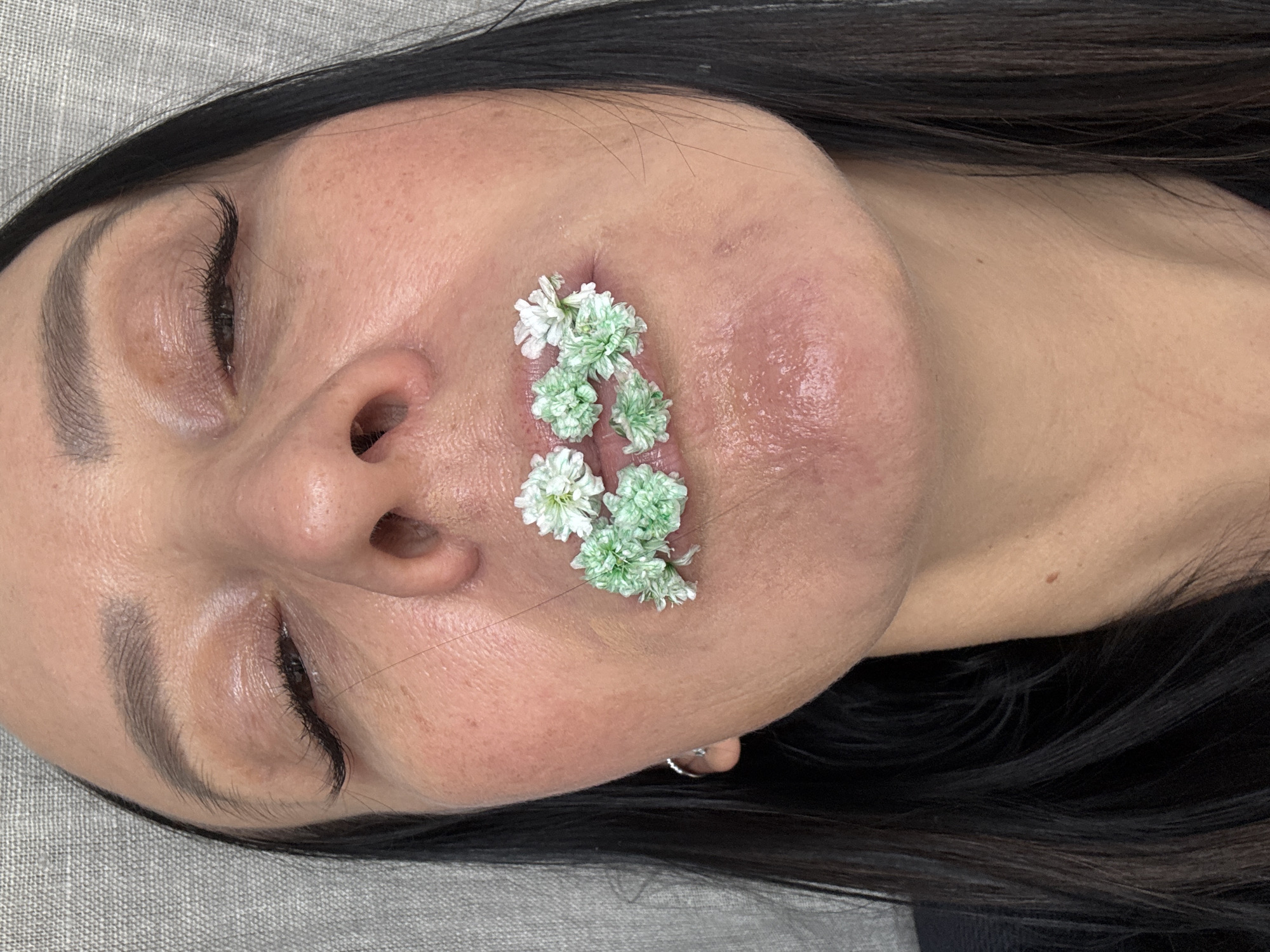 The image displays a close-up, top-down view of a woman's face, resting on a light grey, textured fabric, likely a pillow or treatment bed. Her head is slightly tilted to the side. She has dark hair, which frames the upper and lower portions of her head. Her eyes are closed or angled downwards, revealing well-groomed dark eyebrows and long, dark eyelashes.
The central focus of the image is her mouth, which is adorned with numerous small, white and light green flowers. These delicate florets are arranged along her upper lip and around the corners of her mouth, creating an artistic or decorative effect. Her skin appears to have a light to medium complexion, with some areas around her mouth showing subtle redness or texture. Her lips appear full and well-hydrated, possibly slightly swollen.
The overall mood is serene and calm, emphasized by her closed eyes. There are no other objects or people visible, and no visible text. The scene is likely set indoors, possibly within a beauty salon or photography studio in Chaikovsky, Russia, given the close-up and controlled environment. The activity appears to be a posed shot, possibly for artistic expression or to showcase a beauty treatment or product.