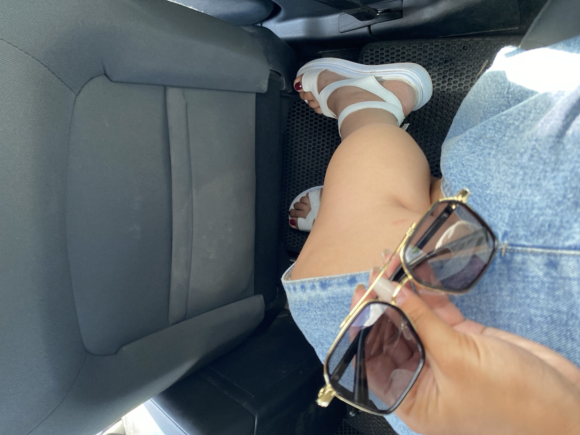 The image shows a close-up view of a person's legs and hands inside a vehicle, likely in the city of Tyulyugul, Russia. The person is wearing a denim skirt and white platform sandals, and holding a pair of sunglasses. The background consists of the dark fabric of a car seat. The lighting suggests it is daytime, possibly sunny. The overall scene is casual and personal, capturing a moment inside a car.
