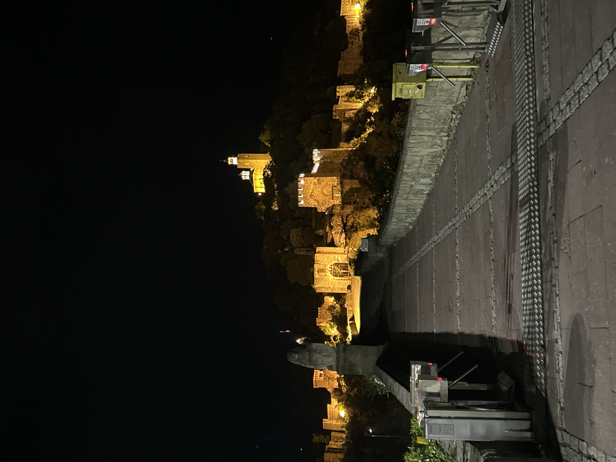The image shows the illuminated Veliko Tarnovo fortress at night. The fortress is built on a hill, with its stone walls and towers visible against the dark sky. Lights illuminate the structure, highlighting its architectural details. A stone walkway with metal railings and tactile paving is in the foreground. Beyond the walkway, a low stone wall separates it from the base of the hill upon which the fortress stands. Below the fortress, a statue can be seen.