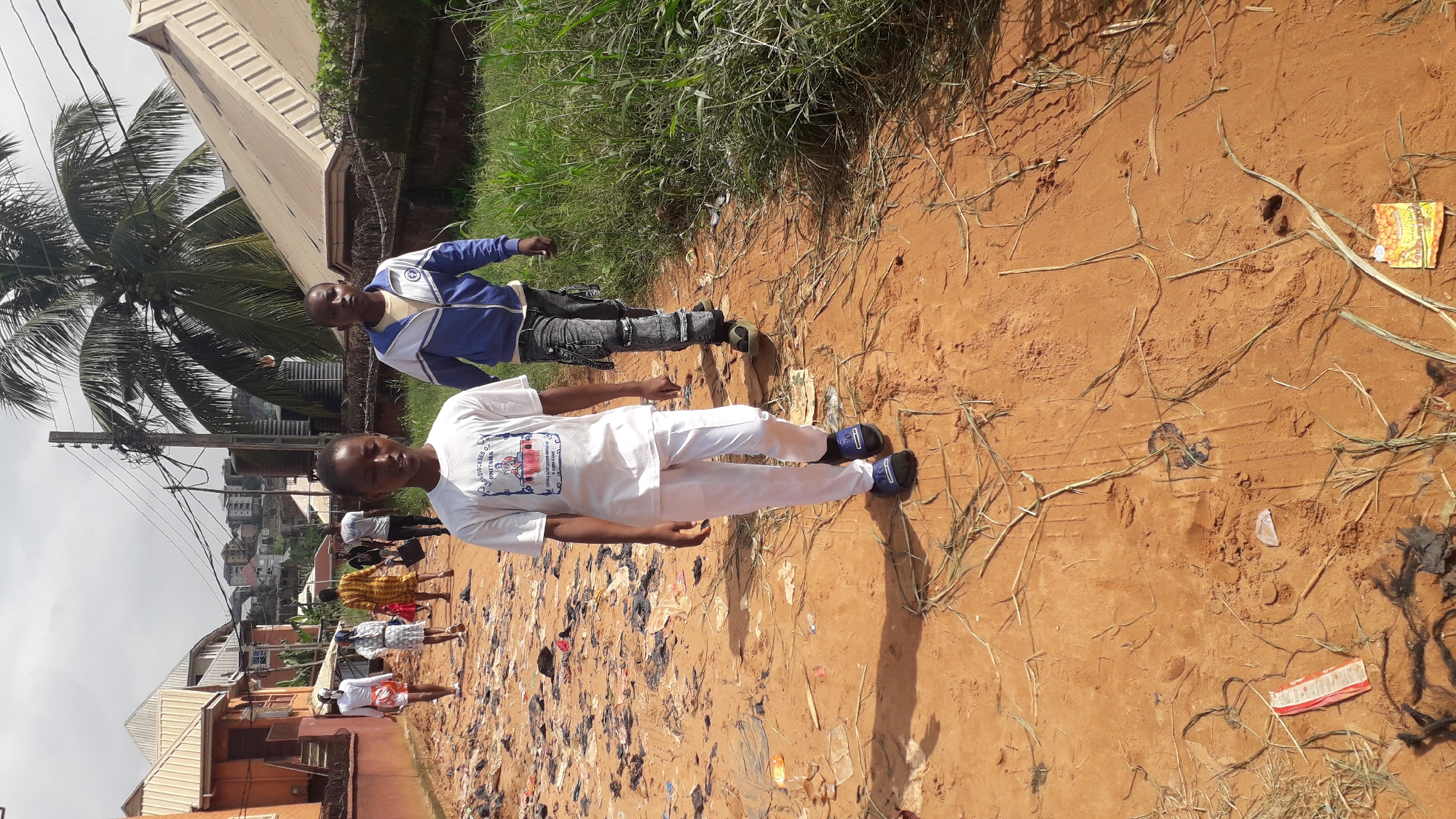 In Nkpor, Nigeria, two young boys walk on a dirt path littered with debris. The boy in the foreground wears a white t-shirt with a graphic and white pants, along with dark blue slides. He is facing slightly to the right, with his head turned toward the camera. Behind him, the second boy wears a blue and white jacket over a yellow shirt and dark, distressed jeans. He is walking towards the right side of the frame.
The environment is characterized by a dusty, reddish-brown path lined with tall, green grass on the right. In the background, several houses with orange and cream-colored walls and corrugated metal roofs are visible. A prominent palm tree with lush green fronds stands behind the boys, and power lines crisscross overhead. The sky is overcast. The overall scene suggests a residential area with some level of disarray due to the scattered trash.