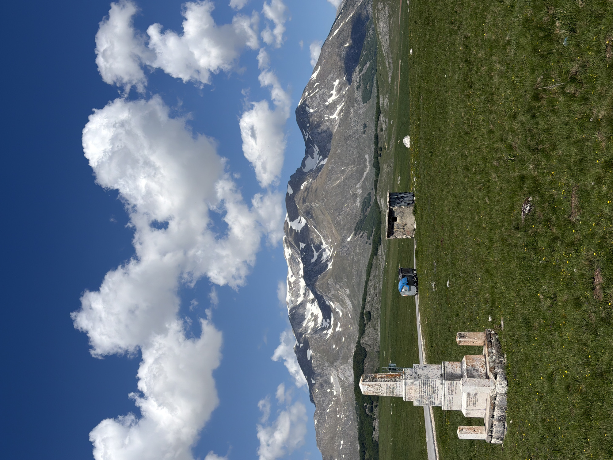 The image shows a mountainous landscape in Novakovići, Montenegro, featuring a memorial monument in the foreground, a grassy field, and a rugged mountain with snow patches extending into the background.
The memorial structure is composed of white stone elements. A small motorized vehicle with blue accents is visible on a path in the middle ground. Behind the path stretches an expanse of green field containing sporadic patches of yellow flowers. The mountain in the background is predominantly gray with areas of snow. A blue sky with scattered white clouds is visible above and to the left of the mountain.