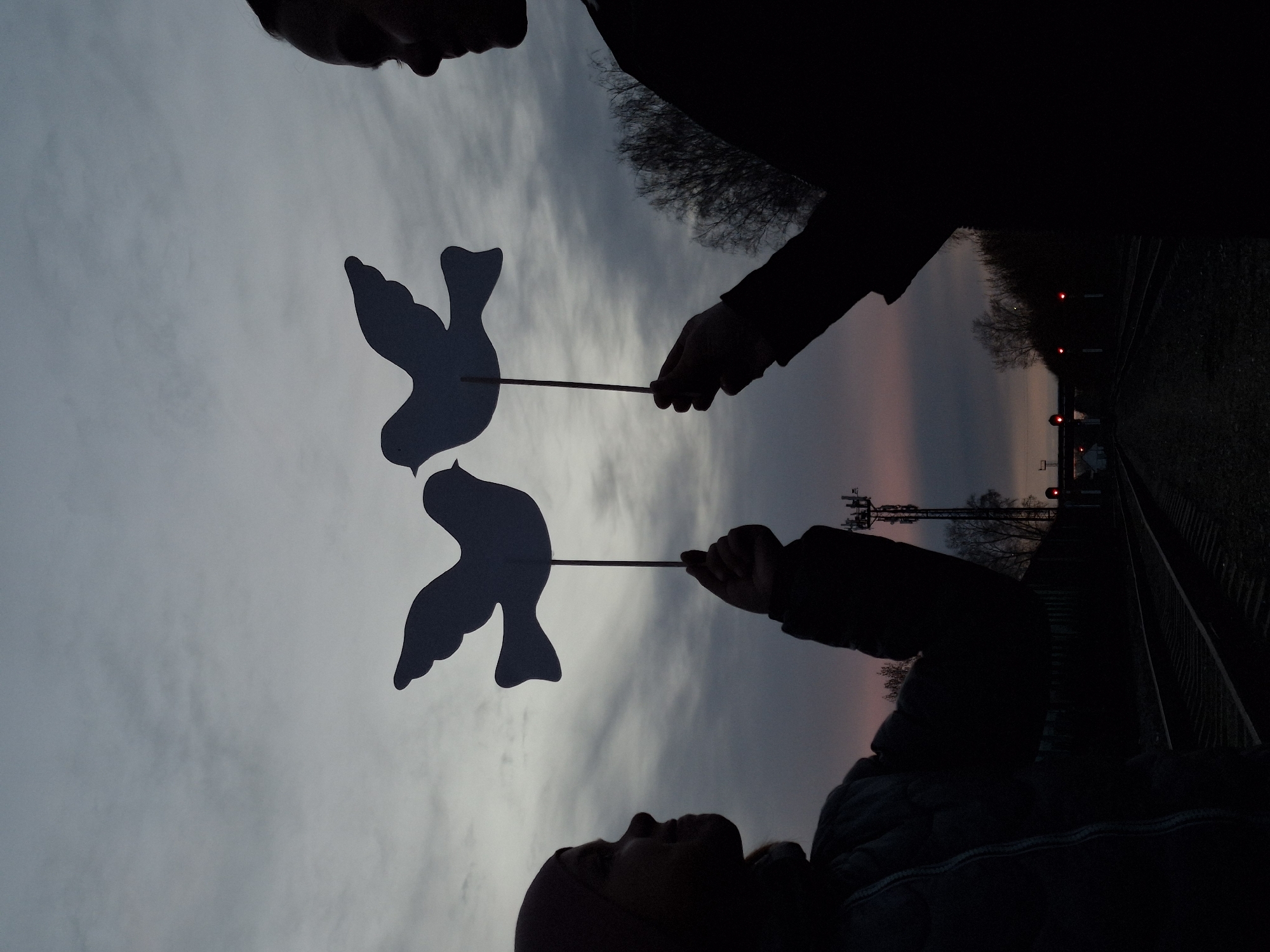Two people are silhouetted against a cloudy sky, holding up cut-out dove shapes on sticks. The sky is a mix of grey and pink, suggesting either dawn or dusk. In the background, a railway track stretches into the distance, with signaling lights visible. Telecommunication towers stand near the tracks. The location is Chaikovsky, Russia, identified by the railway and telecom infrastructure common in the region.