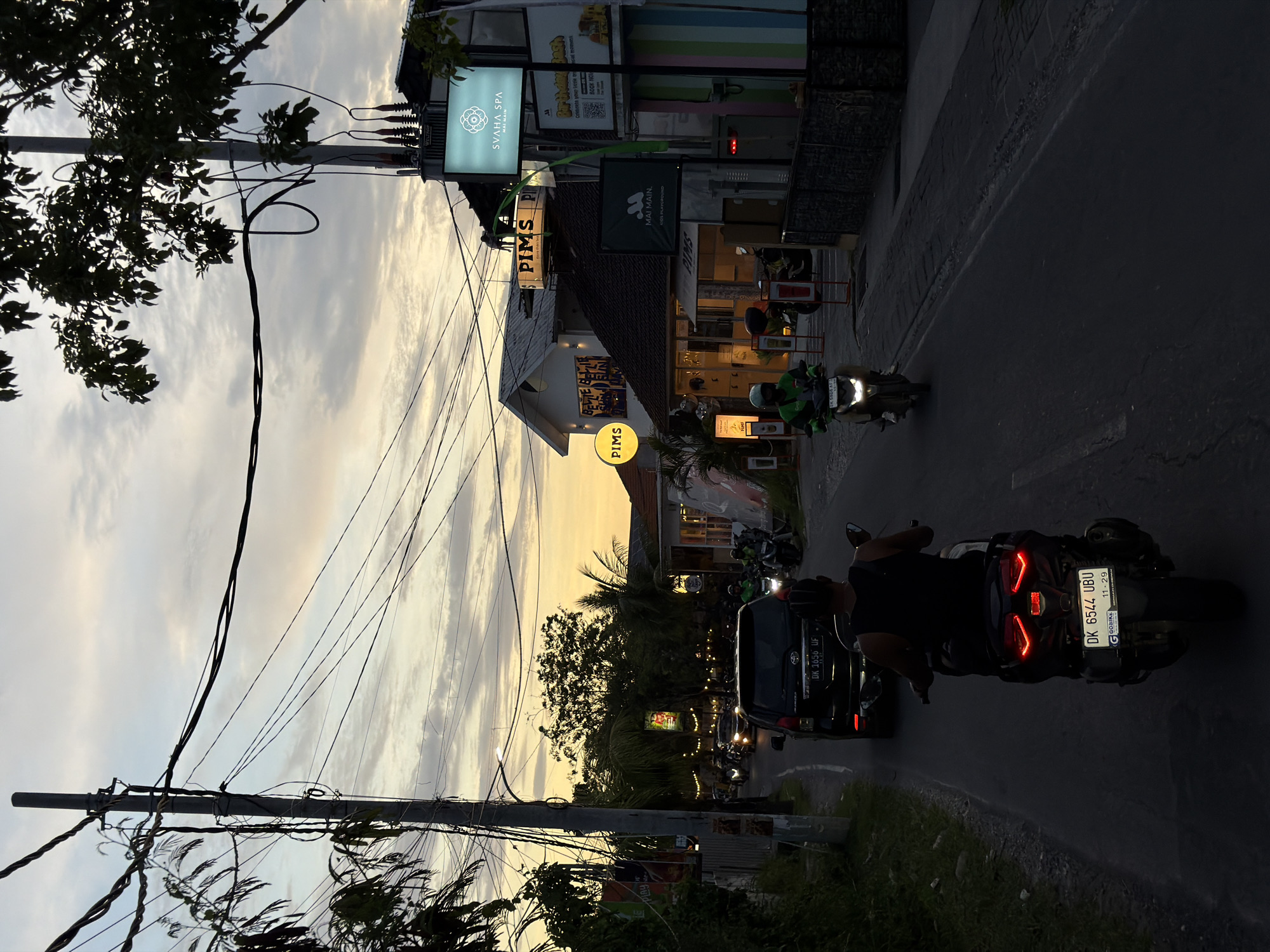 The image, taken from a slightly elevated position, captures a street scene at dusk in North Kuta, Indonesia. The street curves gently to the left, with traffic including motorcycles and a dark-colored car with the license plate "DK 1656 UF". A motorcycle closer to the camera has the license plate "DK 6544 UBU" and a sticker indicating "GGOBIKE" and an expiration date of "11-29".
Buildings line the side of the street, illuminated by artificial light. Signs for "SVAHA SPA", "PIMS", and "MAI MAIN. KIDS PLAYGROUND" are visible. A banner with "Birthday Bash" and "Create your Gittle" is visible above the text "BOOK NOW."
A utility pole with numerous wires stretches across the upper left side of the frame. The sky is filled with clouds, with the setting sun casting a yellow hue. Trees and other vegetation line the edge of the road. People are visible on the sidewalks near the buildings.