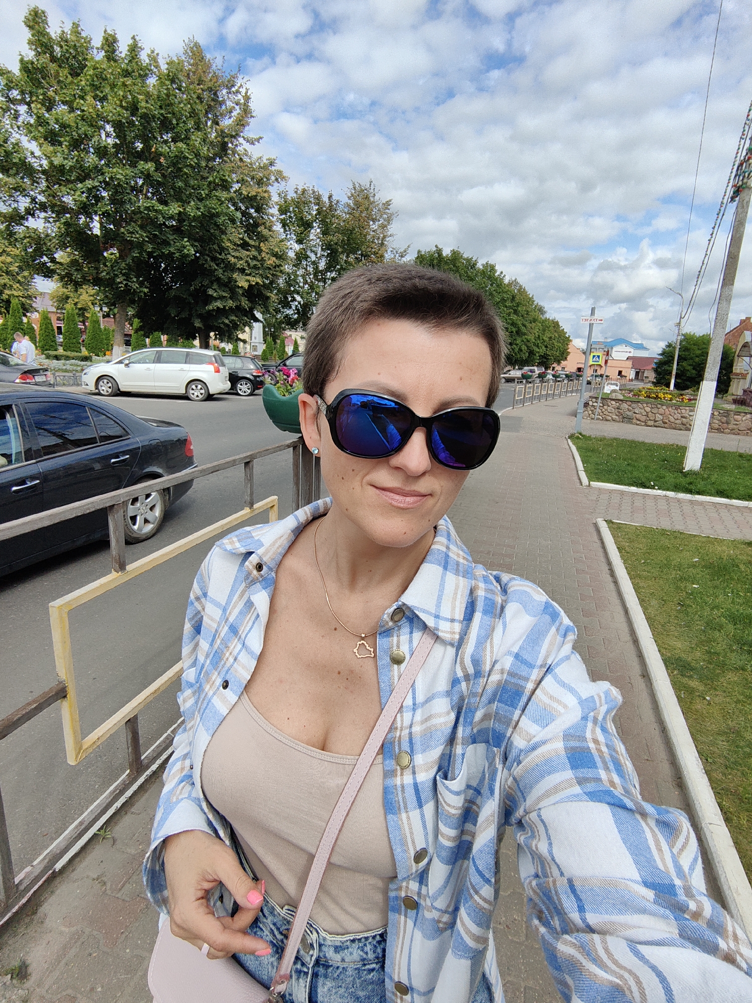 In Pastavy, Belarus, a woman with short brown hair and sunglasses stands on a paved sidewalk, facing the camera. She is wearing a blue and white checkered open shirt over a tan tank top and denim shorts, with a pink cross-body bag. A gold heart-shaped necklace is visible. To her left, a metal railing separates the sidewalk from a street with parked cars, including a black sedan and a white minivan. Trees line the street, and buildings with various architectural styles are visible in the background under a partly cloudy sky.