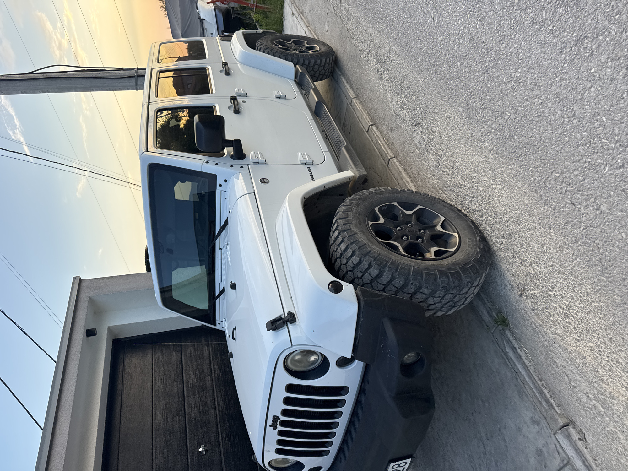 A white four-door Jeep Wrangler is parked on a paved surface next to a garage with a dark wood-grain door. The Jeep is positioned at an angle, with the front left of the vehicle closer to the camera. The sky is a clear blue with some wispy clouds, and the sun appears to be setting, casting a warm glow on the scene. In the background, there is a power pole and some vegetation. The image was taken in Burtaiši, Montenegro.