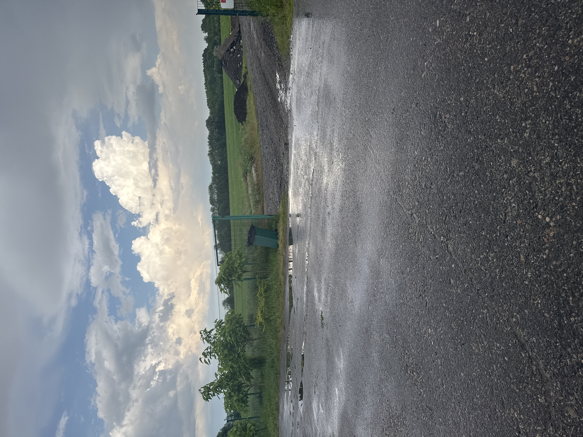 The image depicts an outdoor scene with a focus on a wet asphalt surface in the foreground. Puddles of water are scattered across the pavement, suggesting recent rainfall. Beyond the asphalt, a green metal fence stretches across the middle ground. To the left of the fence, several young trees with lush green foliage are planted in a grassy area. A green trash can stands near the fence. The background features a wide expanse of green fields, with a treeline visible in the distance. The sky above is filled with dramatic clouds, with patches of blue sky showing through. A pile of dark material, possibly dirt or gravel, is visible in the middle ground on the right side. The overall impression is of a somewhat overcast day in a semi-rural or industrial area.