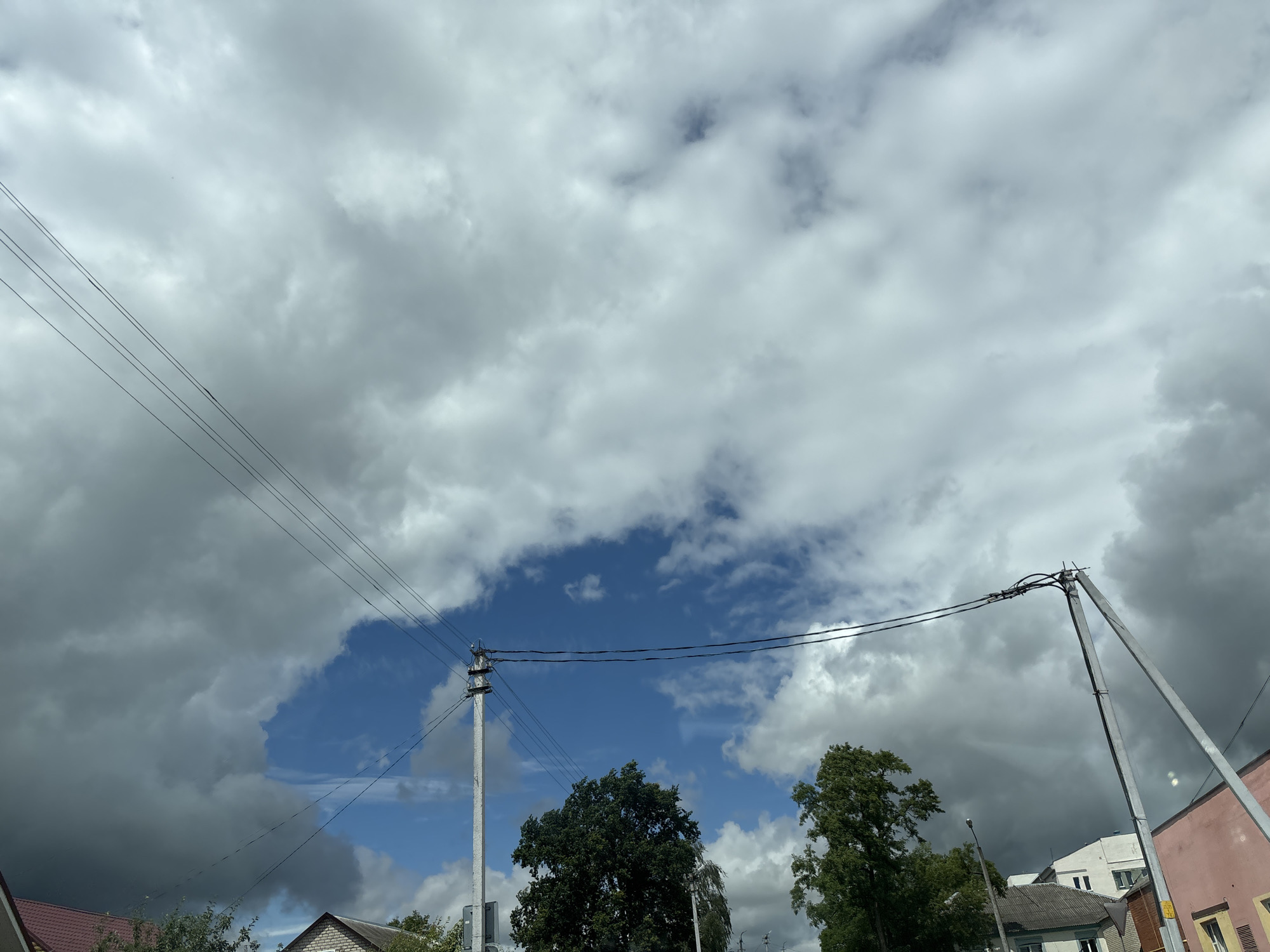 The image captures a suburban scene in the vicinity of Капыльскі сельскі Савет, Belarus, under a dramatic sky. The upper portion of the frame is dominated by a heavy, overcast sky with large, dark storm clouds. A break in the clouds reveals a patch of clear blue sky. Several telephone poles with multiple wires stretch across the frame, connecting various points and contributing to the utilitarian aesthetic of the area. In the lower section, the tops of houses are visible, characterized by pitched roofs. Trees with dense green foliage are interspersed between the buildings, adding a touch of nature to the urban landscape. The overall impression is one of an impending or recent weather event, with the stark contrast between the dark clouds and the blue sky being a prominent feature.