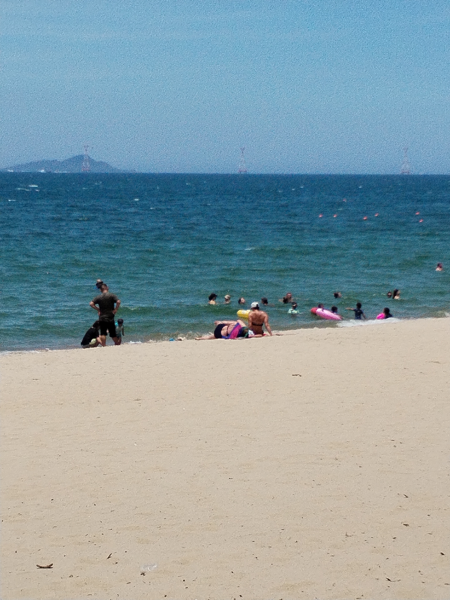 The image depicts a beach scene in Nha Trang Ward, Vietnam, with a wide expanse of sand in the foreground and the ocean stretching into the background under a clear blue sky. Several people are visible on the beach and in the water. On the sand, a man wearing a dark t-shirt and shorts stands with his hands on his hips, facing the water, with two children near him. To the right, two individuals are lying on the sand, one wearing a bikini and the other in swimwear. In the water, a group of people are swimming and wading, with some using inflatable floats, including a pink one. In the distance, the horizon shows the sea meeting the sky, with hints of landmasses and structures, possibly towers or buildings, on the far horizon. The water is a deep blue with visible waves.