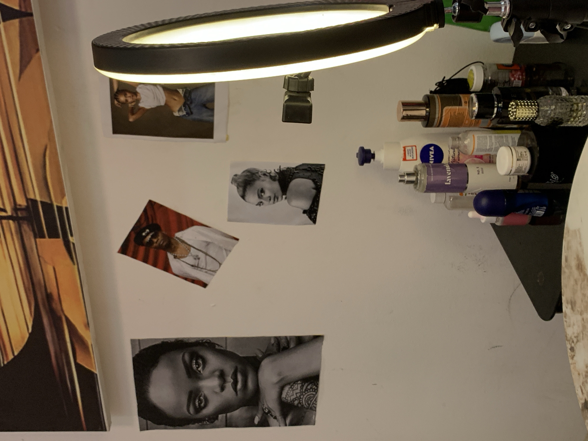The scene is an interior room located in Lagos, Nigeria. A predominantly white wall serves as the background, displaying an abstract, horizontally oriented painting in shades of gold and dark brown/black at the top. Below the painting, four photographic prints are affixed to the wall. From left to right: a black and white portrait of recording artist Rihanna, depicted looking directly forward with a tattoo visible on her right arm; an angled color photograph of an unidentified male subject wearing sunglasses, a dark cap, and multiple chain necklaces; a black and white portrait of recording artist Beyoncé, looking over her left shoulder; and a color photograph of an unidentified female subject in a white crop top and jeans, posing with hands behind her head, partially obscured by foreground equipment. To the right, a large, circular LED ring light, emitting a warm yellow glow, is positioned on a dark stand with an attached camera mount, facing towards the left. In the lower portion of the image, a glossy black surface, presumably a dressing table or shelf, supports a collection of personal care products. Notable items include a white NIVEA brand pump lotion bottle, a purple-labeled bottle containing 'lavender' product, various perfume bottles of different shapes and sizes (including one with a rose-gold cap and a clear patterned bottle), a dark blue roll-on dispenser, and several smaller jars and containers (one white with a gold lid, one dark, and a transparent jar with a yellow lid containing small edibles). The wall exhibits minor surface marks and texture variations. The overall illumination is artificial.