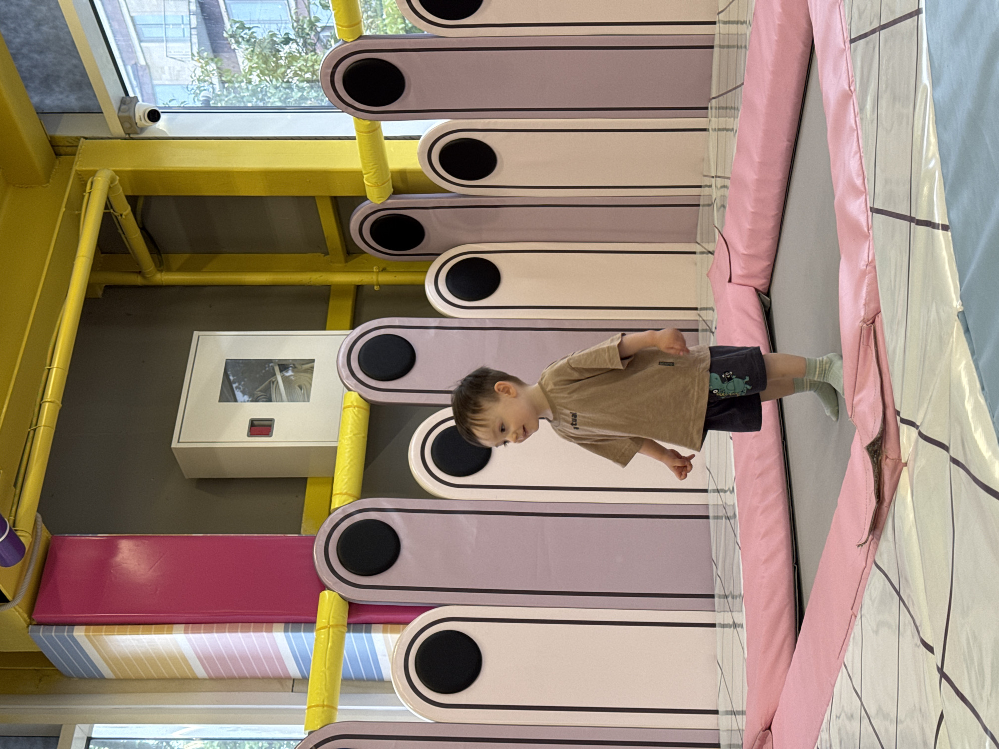 A child stands on a trampoline in an indoor play area. The play area features cushioned steps or platforms in pink, purple, white, and yellow. The area is well-lit by natural light coming from a window. There are no indications in the photograph that indicate its location as Sochi, Russia.