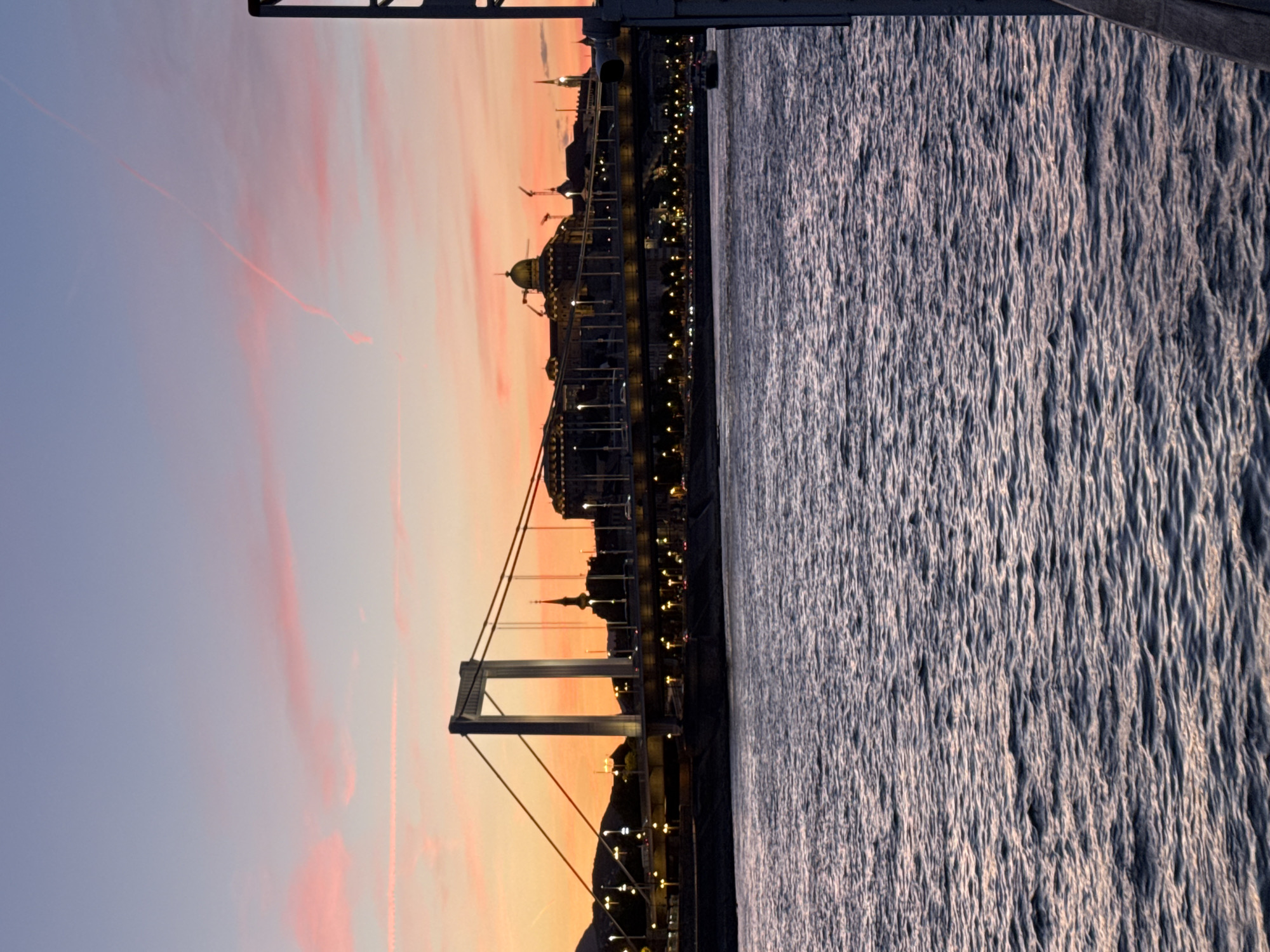 The image presents a serene panoramic view of Budapest, Hungary, during either sunrise or sunset, looking across the Danube River towards the city skyline. The sky displays a beautiful gradient, transitioning from a soft, pale blue at the top to warm orange, pink, and red hues closer to the horizon, with delicate, wispy clouds streaking across.
The calm waters of the Danube River occupy the foreground and mid-ground, reflecting the ambient light and forming a textured surface of gentle ripples. Stretching across the river, a prominent white suspension bridge, likely the Elisabeth Bridge, is a central feature. Its strong pylons and cables are visible, creating a striking silhouette against the colorful sky, yet also subtly illuminated.
On the far bank, the city's architecture defines the skyline. Numerous buildings and structures are visible, some standing as dark silhouettes while others are adorned with twinkling artificial lights that punctuate the evening or early morning scene. Distinct architectural elements such as a large building with a dome and several spires characteristic of Budapest are discernible. The overall setting is urban and majestic, showcasing the city's beauty bathed in the soft glow of dusk or dawn. No individuals or visible text are present in the image.