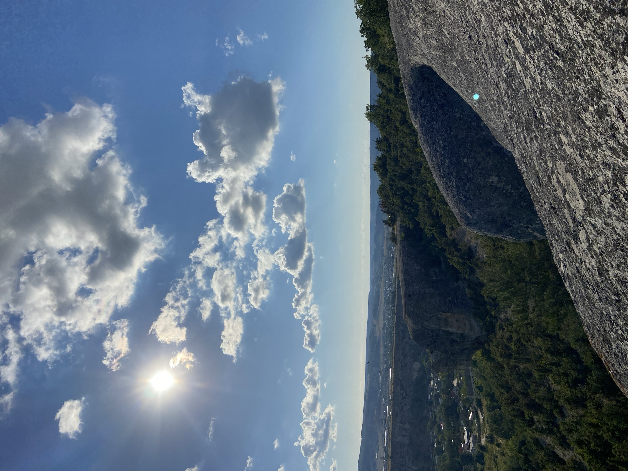A scenic view overlooks a valley with a village nestled within it, under a bright blue sky dotted with white and gray clouds. The sun shines brightly in the upper left corner, casting a lens flare. In the foreground, a rough, gray rock formation dominates the lower right portion of the frame, with its textured surface catching the light. The middle ground features a steep, rocky cliff face covered in green trees and vegetation. Beyond the cliff, the valley opens up, revealing houses and buildings of a settlement. Further in the distance, rolling hills and mountains form the horizon line, all bathed in the soft light of what appears to be late afternoon. A single bird is visible in flight in the middle distance. The image was taken in the village of Krasnomakskoye in Ukraine.