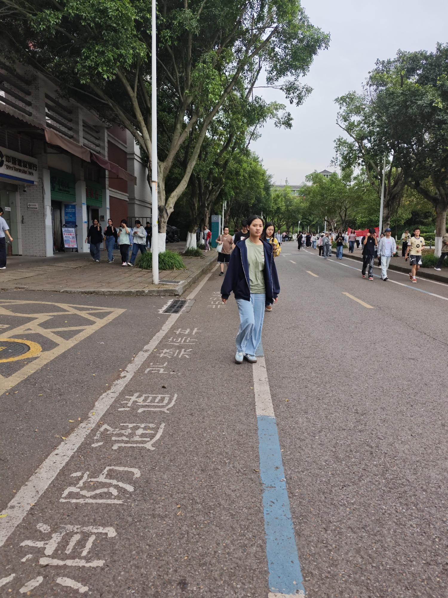 The image captures a street scene in what appears to be a university campus setting in Chongqing High-tech Zone, China. A young woman dressed in a dark blue jacket over a light green top, light blue jeans, and white sneakers is walking towards the camera along a road lane marked with a blue stripe. To the left of the woman, on the side of the road, there is a pedestrian walkway with white painted Chinese characters. Further to the left is a building with retail fronts and several people walking along the sidewalk.
Behind the woman and along the right side of the road, a diverse group of people are also walking in the same direction, seemingly participating in a larger event or gathering. Tall trees line both sides of the road, creating a canopy overhead. The sky is overcast.