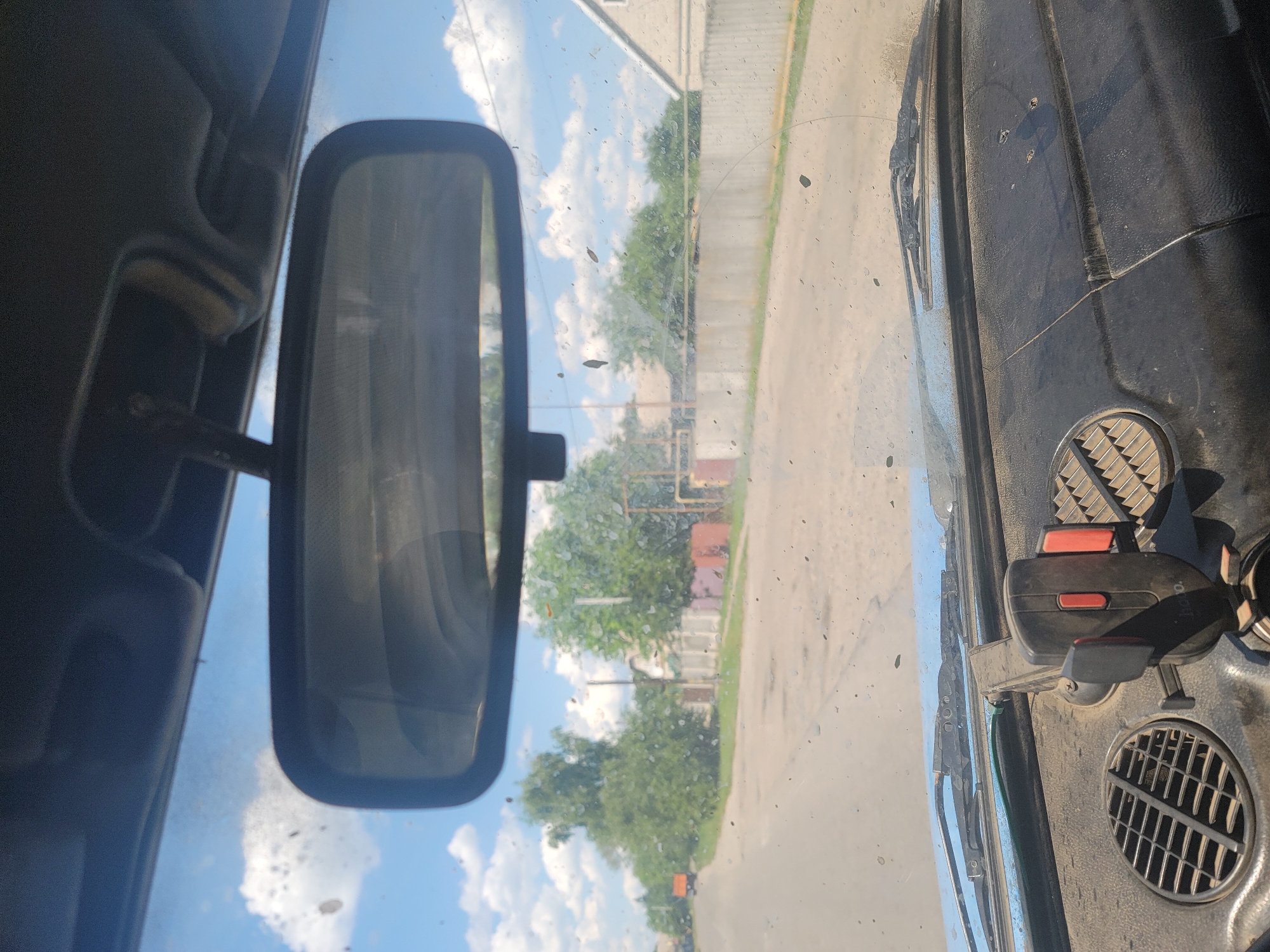 The image is a point-of-view shot from inside a vehicle, looking forward through a dusty windshield. The scene appears to be a rural road in Pereshchepyne, Ukraine, under a bright, cloudy sky. The dashboard is visible in the foreground, cluttered with a phone mount, air vents, and general wear and tear. The windshield itself is smudged with dirt and small debris.
Beyond the windshield, a paved road stretches into the distance, flanked by trees and houses. A red truck is visible in the far left, driving away from the camera. Fences and various structures are present along the roadside, suggesting a residential area. The overall impression is that of a mundane drive through a village on a clear day. There are no people clearly visible in the image.