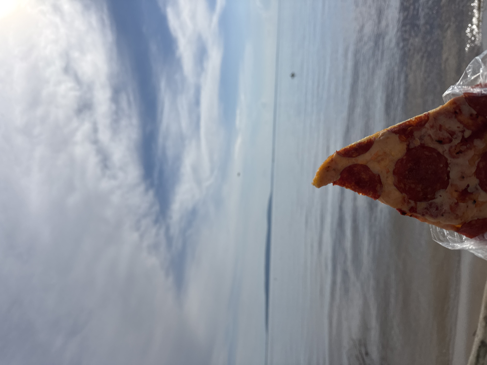 This media file shows a slice of pepperoni pizza held up against a cloudy sky and a body of water. The pizza is in the foreground, taking up the right side of the frame. It appears to be a typical slice with cheese and pepperoni. The background consists of a vast sky filled with white and gray clouds, with patches of blue visible. Below the sky is a calm body of water, likely a lake or the ocean, reflecting the sky. The overall scene suggests an outdoor setting, possibly a beach or a pier. The lighting indicates it could be daytime, but the overcast sky suggests possibly diffused sunlight. There is no visible text in the image. The image is taken from a low angle, looking up towards the sky and the pizza.