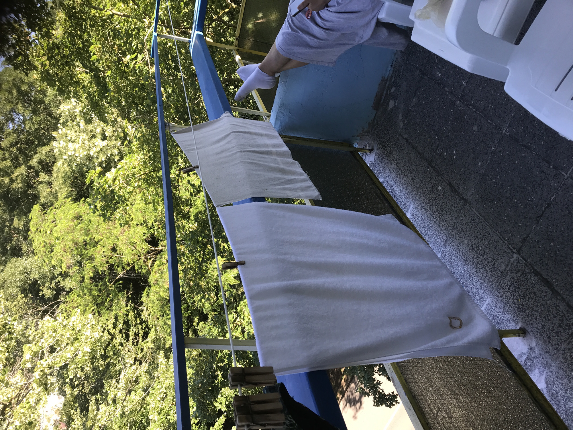 A person, wearing white socks and a light gray robe, is relaxing on a balcony in Varna, Bulgaria. They are reclined with their legs extended, appearing to be holding a smartphone in their right hand. Two white towels are hung on a clothesline strung across the balcony, secured with wooden clothespins. The balcony overlooks a dense area of lush green trees. A white plastic chair is visible on the right side of the frame. The balcony floor is paved with dark, speckled tiles, and the railing is painted blue. The scene is bathed in sunlight, suggesting it is daytime.