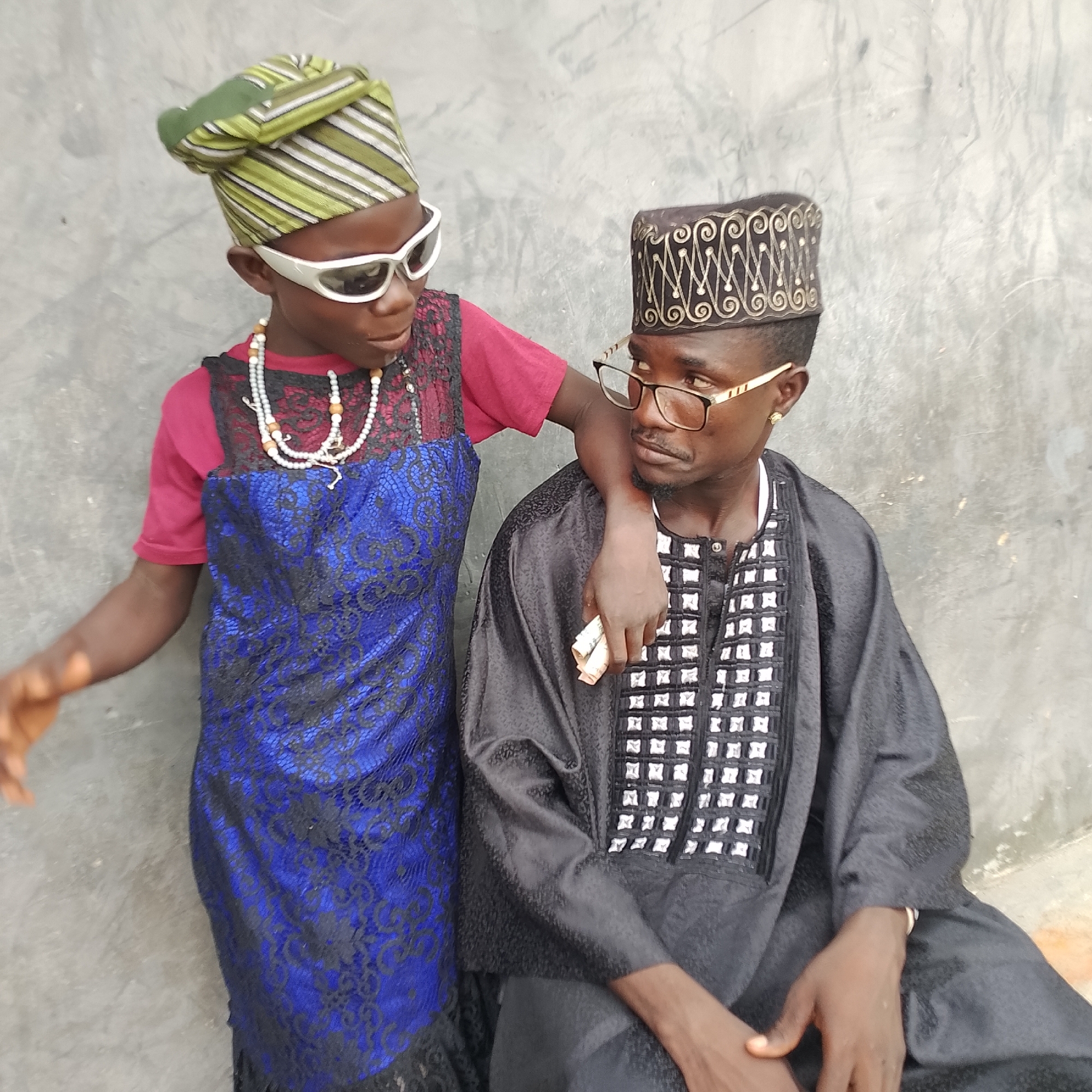 Two individuals, one appearing to be a child and the other an adult male, are posing for a photograph against a textured, grey wall. The child, on the left, wears a maroon shirt under a blue lace dress, white sunglasses, and a green and white striped headwrap. A white beaded necklace is visible. The child's left arm is draped over the adult male's shoulder. The adult male, on the right, is wearing a black cap with intricate gold embroidery, glasses with gold frames, and a black, embroidered garment. He is holding a few folded banknotes in his right hand. Both individuals are looking towards the camera or slightly off to the side. The environment suggests an outdoor urban setting, possibly in Ado, Nigeria.