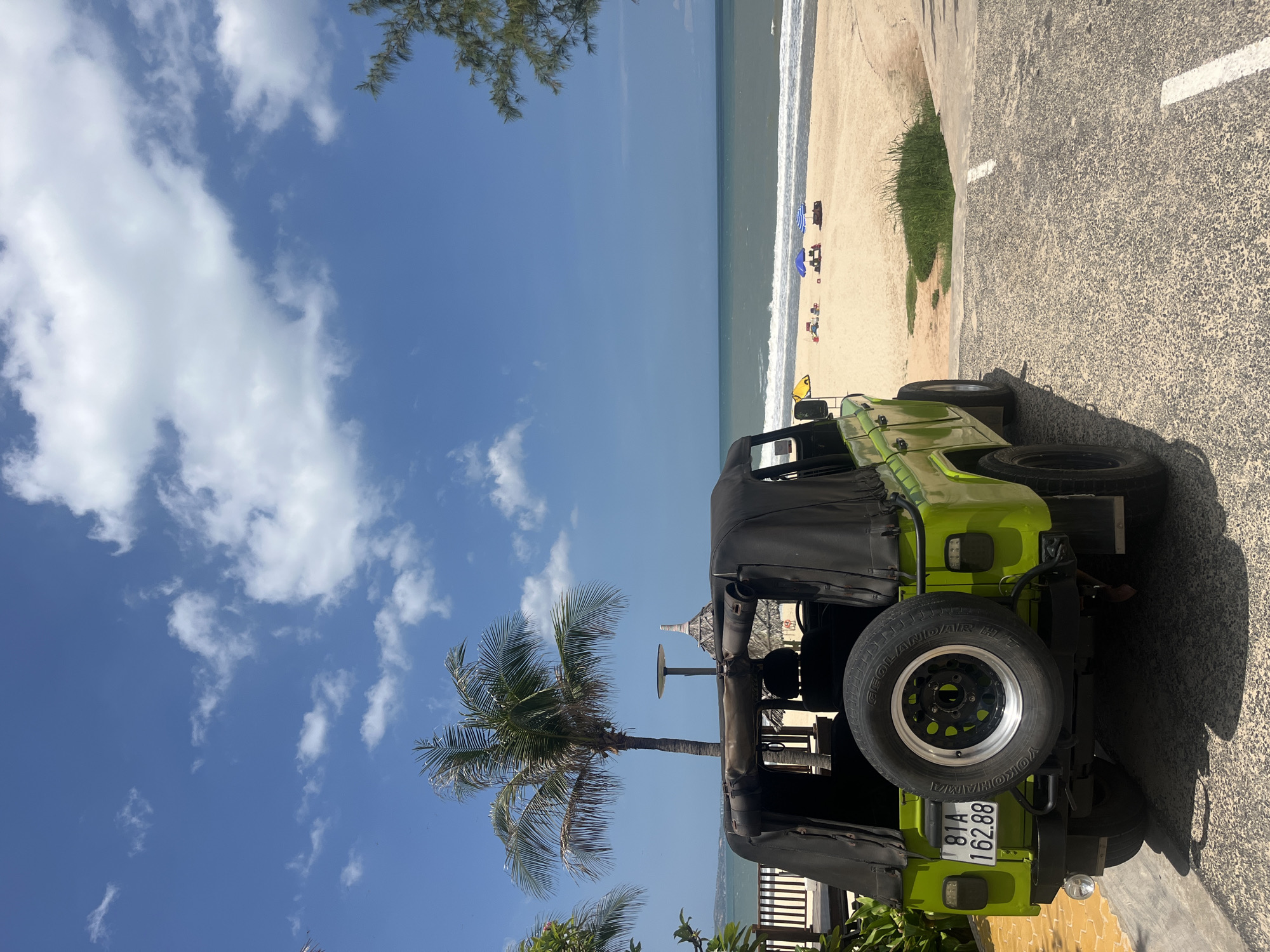 This is an outdoor, eye-level, horizontal medium shot of a bright green Jeep parked on a paved surface next to a sandy beach and the ocean. The Jeep is the primary subject, with its top down and a spare tire mounted on the back. The beach is visible in the background, with a few beach umbrellas and chairs set up. Several people are scattered on the beach, and the ocean waves are breaking on the shore. A palm tree is in the foreground on the left, with its fronds extending towards the Jeep. The sky is a clear blue with some white clouds. The sun appears to be high in the sky, suggesting it is daytime, likely midday. The weather appears to be sunny and pleasant, suitable for a beach outing. The license plate on the Jeep reads "81A 162.88". The scene evokes a sense of relaxation and vacation.