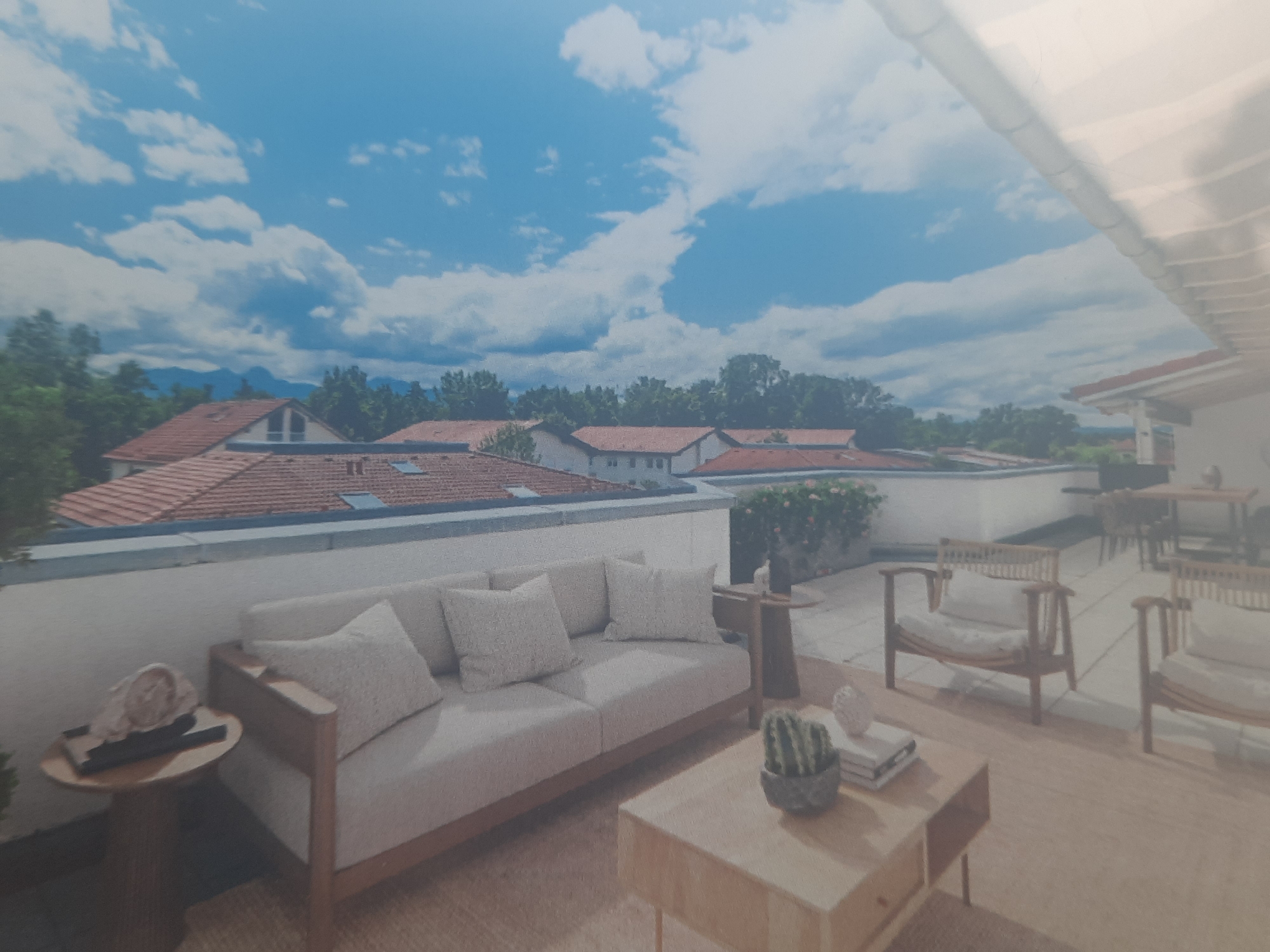The image depicts a bright, airy outdoor living space, likely a balcony or terrace, overlooking a scenic view. The foreground is furnished with a comfortable-looking light-colored sofa adorned with several matching throw pillows. To the left of the sofa, a small, round wooden side table holds a decorative shell and two dark rectangular objects, possibly remotes. A low, rectangular wooden coffee table sits in front of the sofa, displaying a potted cactus and a stack of books. In the mid-ground, two wooden chairs with light cushions are positioned on the terrace. Further back, a small wooden table supports a decorative item. The background reveals a rooftop with red tiles, green trees, and mountains in the distance under a vibrant blue sky filled with fluffy white clouds. The overall impression is one of a tranquil, modern outdoor living area. While the image itself does not contain any direct clues indicating the specific location in Rosenheim, Germany, it portrays a scene that could be found in a residential area of the city, perhaps an apartment building or a house with a well-appointed outdoor space.