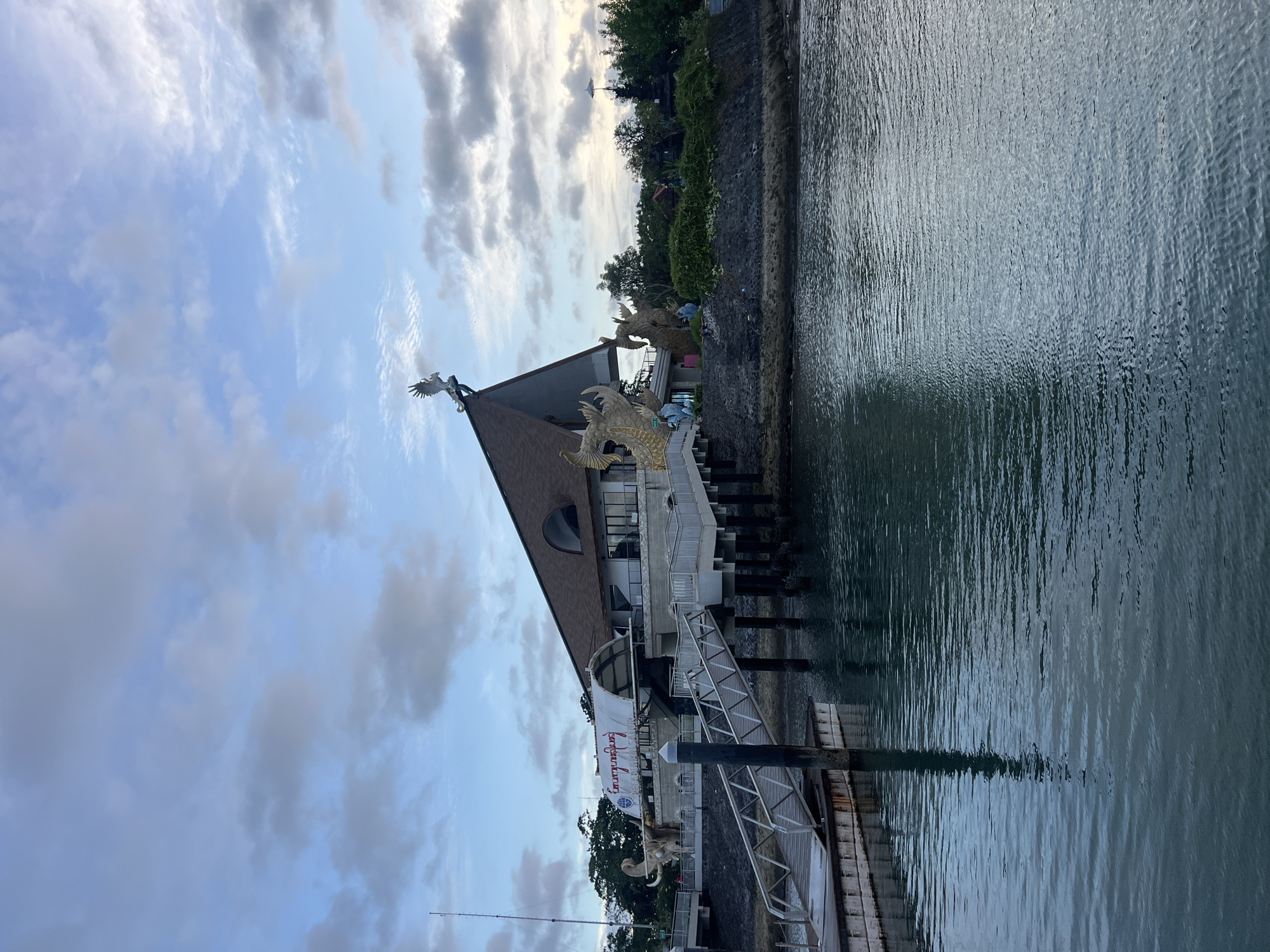 The image, oriented vertically, captures a distinctive building situated by a body of water under a partly cloudy sky. The central subject is a building with a prominent, dark-colored peaked roof. Perched on the roof's apex is a white sculpture of a bird, likely a Garuda, with outstretched wings. Along the building's facade and closer to the water, several large, ornate golden dragon-like sculptures are visible. A significant portion of the image is occupied by a dark, rippling body of water, possibly a river or bay.
A metal walkway or ramp extends towards the water, supported by dark pilings, with additional wooden pilings partially submerged. A stone embankment, lush with green trees and bushes, forms the background behind the building and along the water's edge. The sky displays scattered white and grey clouds against a light blue backdrop, suggesting either late afternoon or early evening lighting. A thin line, possibly a fishing rod, can be seen extending into the lower left corner. A white banner hanging from the building clearly displays the text "Saraswati Luxury." The architectural style and mythological sculptures are consistent with the cultural motifs found in Denpasar, Indonesia.