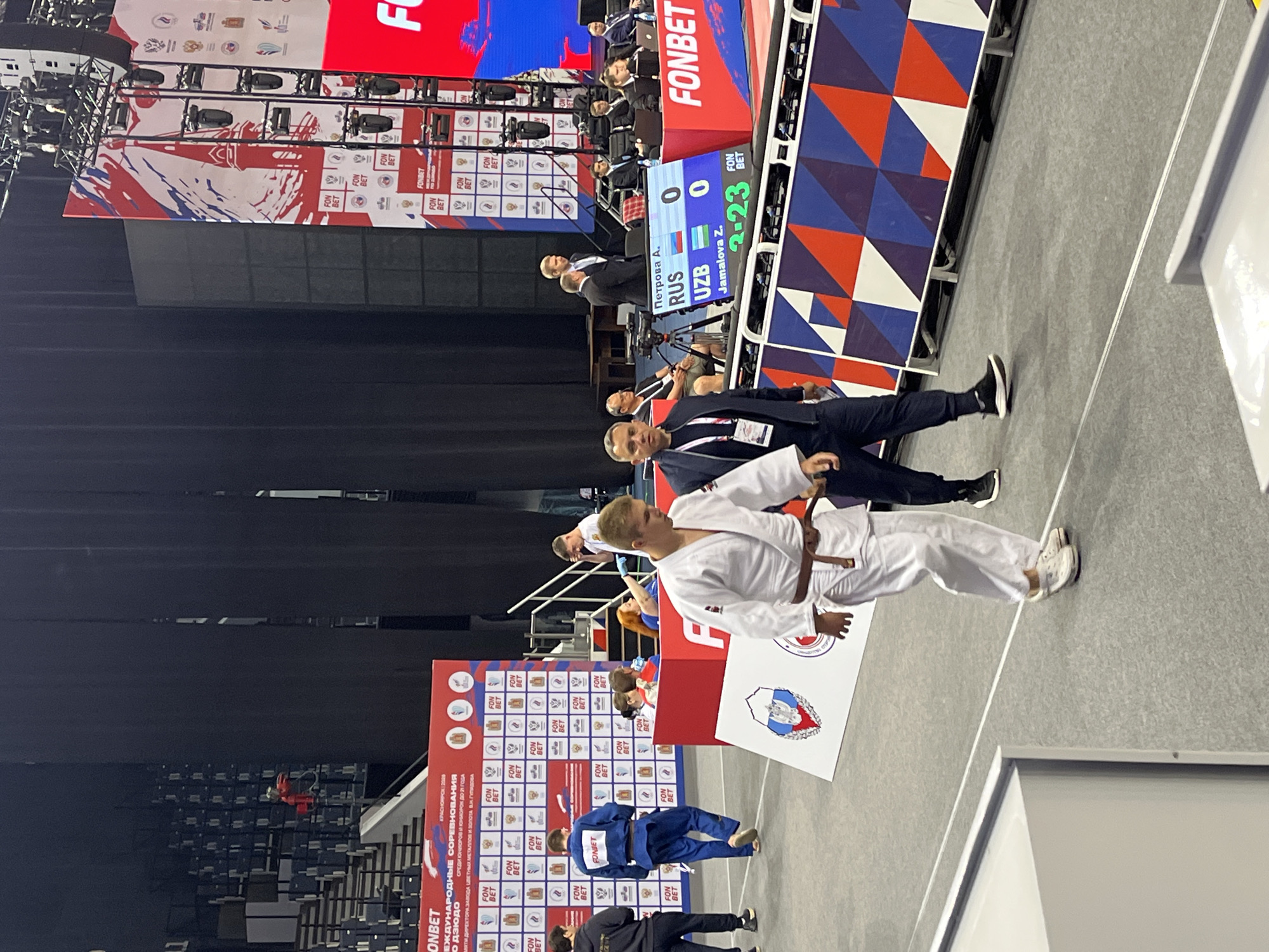 The image shows a judo match in progress at an indoor arena. The scene is a professional sporting event, likely a championship given the scale of the setup. The judoka are wearing traditional white gis with brown belts. One judoka, in the foreground, is in focus, walking forward with his arms slightly bent. Behind him, another judoka, wearing a blue gi with "FONBET" written on the back, is also walking. A scoreboard in the background displays names and scores, indicating the ongoing competition. Several officials and spectators are visible in the background, some seated and others standing. The environment includes stadium seating, large screens displaying event branding, and professional lighting rigs. The branding on the screen and banners indicates the event is sponsored by "FONBET" and is being held in Krasnoyarsk, Russia, as suggested by the text "KPACHOHPCK | 2020".