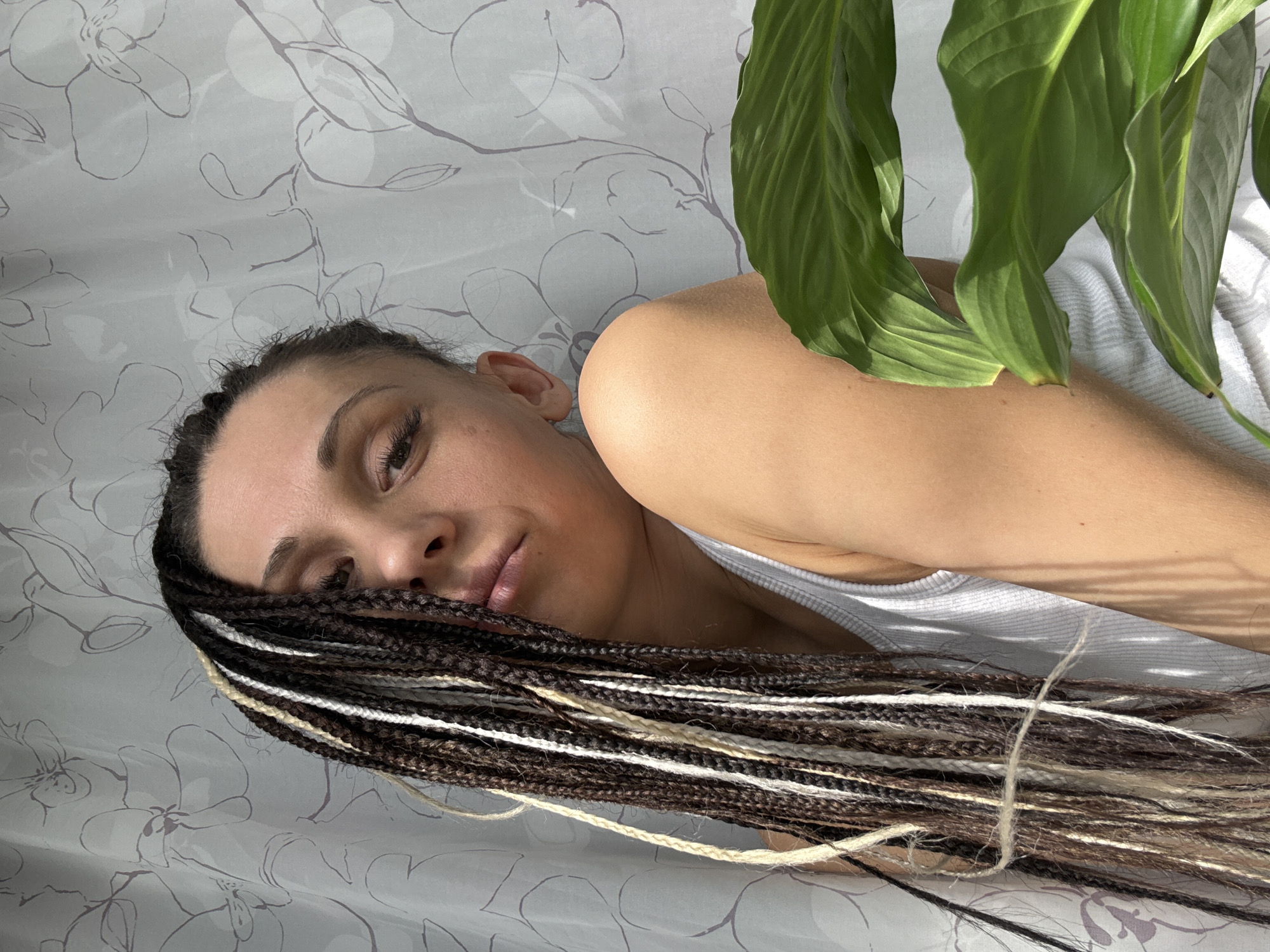 A woman with long, multi-colored braids is lying down, partially obscured by large green leaves. She is wearing a white tank top. The background is a light gray patterned surface. The scene is indoors and the location cannot be determined from the image alone, nor can it be determined if the image was taken in Rusanovo, Russia.