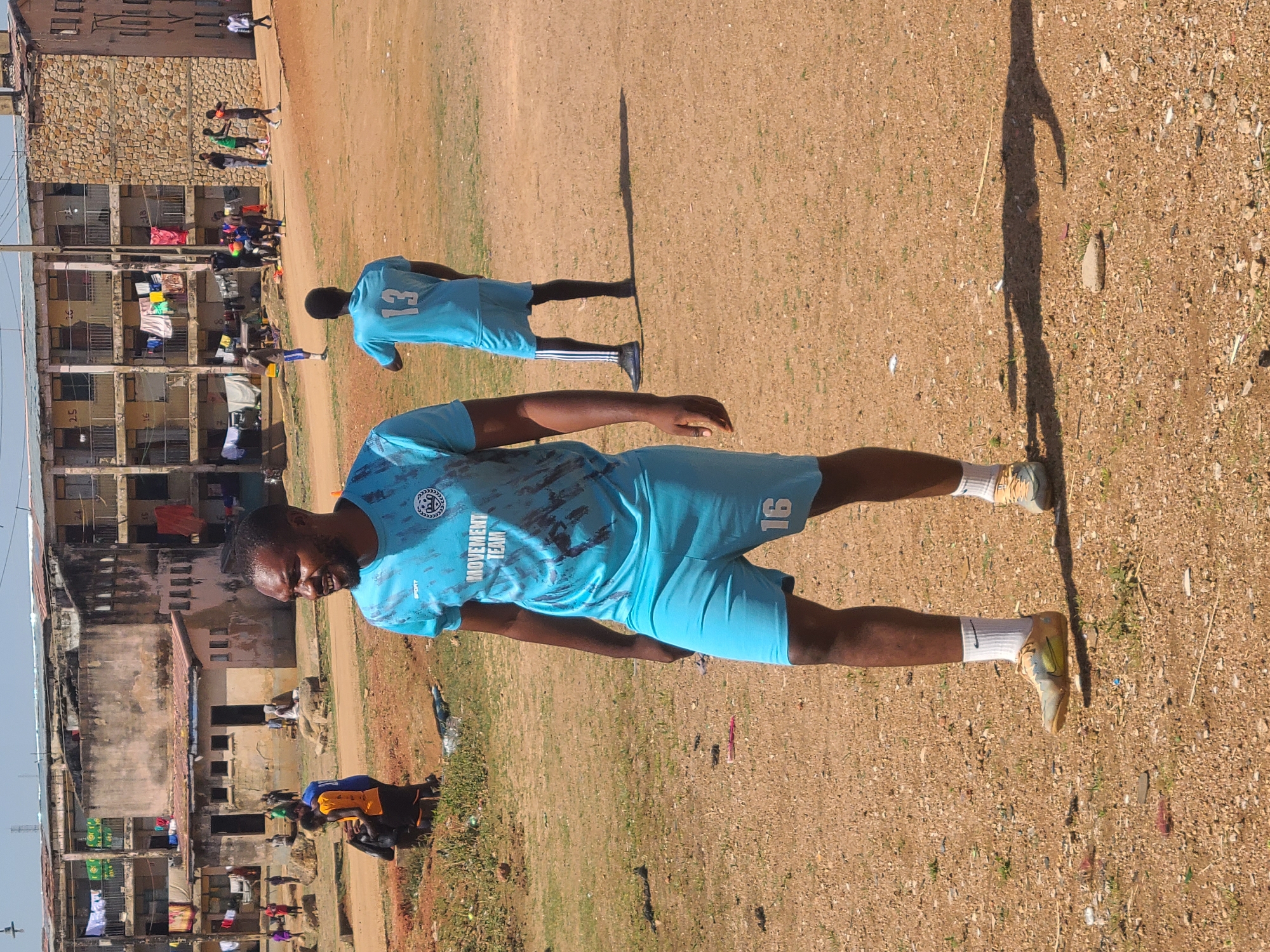 A man is standing on a dirt field in Unknown, Unknown, wearing a blue athletic jersey and shorts. The jersey has the number 16 on the shorts and text that says "MOVEMENT TEAM" on the front of the shirt. Another person is standing behind the man with the jersey, wearing a blue shirt with the number 13. In the background there are buildings and a group of people playing.