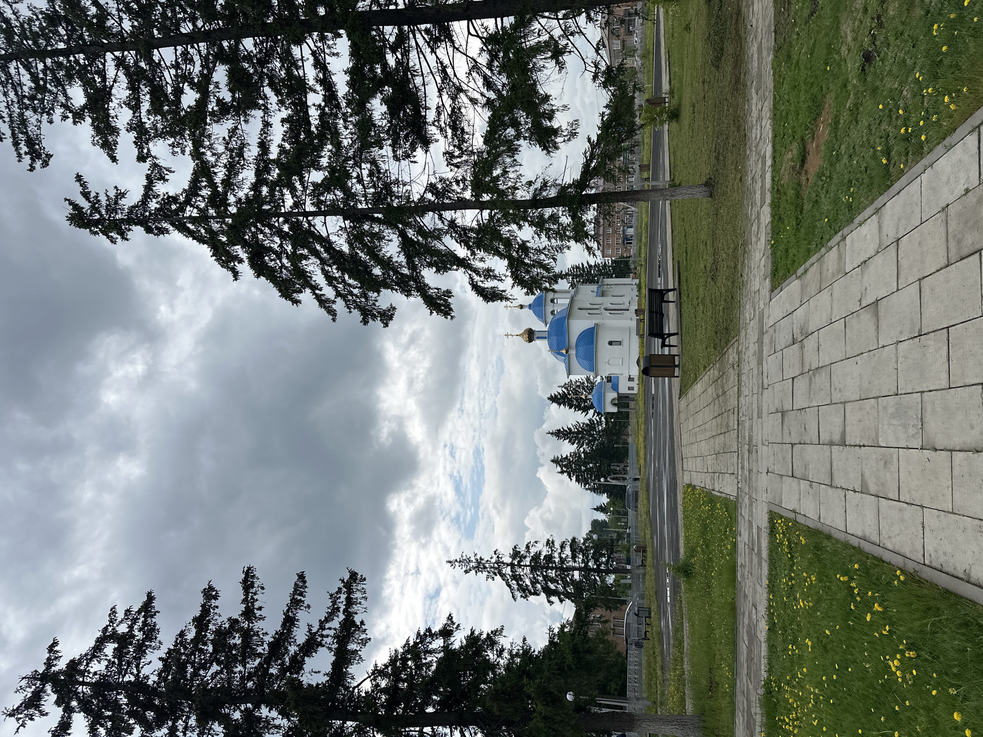 The image shows an overcast day in Dzerzhinskoye, Russia, with a clear view of a white church with blue domes and a golden cross. The church is located in the background, beyond a tree-lined street with a few cars. In the foreground, a paved walkway leads towards the church, bordered by grassy areas with scattered yellow wildflowers. Two benches are visible on the grass near the walkway, and two trash receptacles are situated near the church. Tall evergreen trees frame the scene on either side. The sky is filled with heavy, gray clouds, suggesting a potential for rain.
