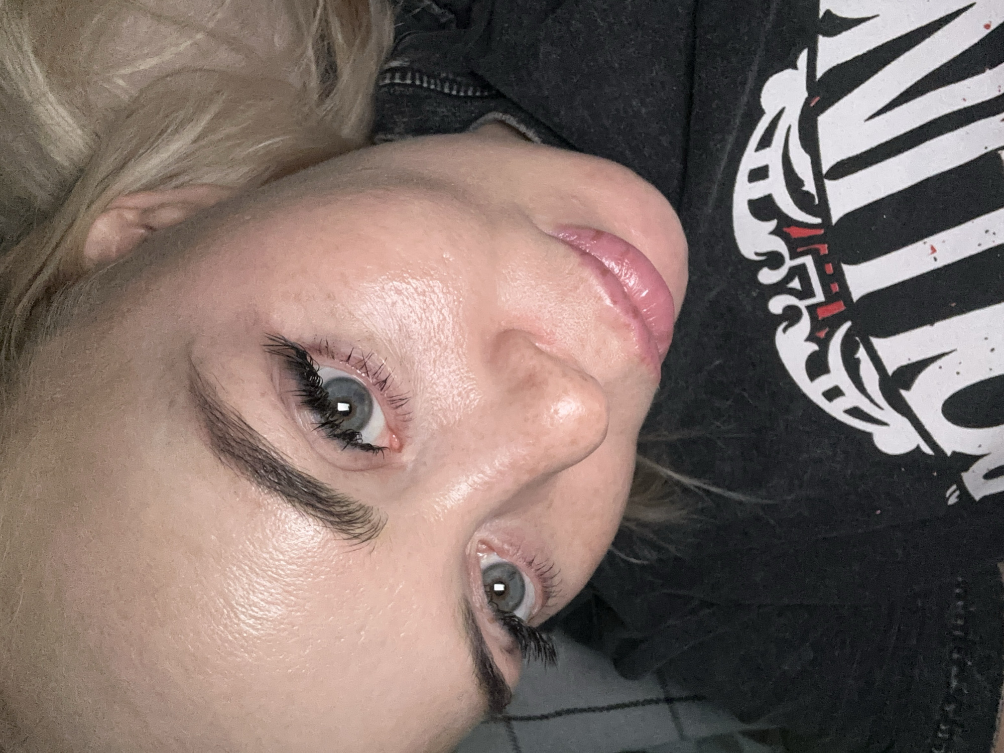 A close-up, high-angle shot features the face of a person, likely female, lying down and looking upwards towards the camera. The subject has blonde hair, visible on the left side of the frame, and a pale complexion with visible pores and a somewhat shiny or dewy appearance on the forehead, nose, and chin. Their eyes are light blue or grey, enhanced by long, dark eyelashes (possibly extensions or mascara) and well-defined, dark eyebrows. The lips are a natural pink tone with a slight sheen.
The person is wearing a dark-colored garment, appearing to be a t-shirt or hoodie, visible on the right side of the frame and beneath their head. On the right side of the garment, there is a prominent graphic design in white and red, which depicts stylized skeletal ribs and other abstract, tribal-like shapes. No clear, legible text is identifiable within this graphic.
The lighting appears to be artificial, possibly a flash or overhead light, which causes bright reflections in the eyes. The background is dark and indistinct, with a faint suggestion of a plaid or checked pattern in the bottom left corner, likely a blanket or pillow. The person's expression is neutral.
