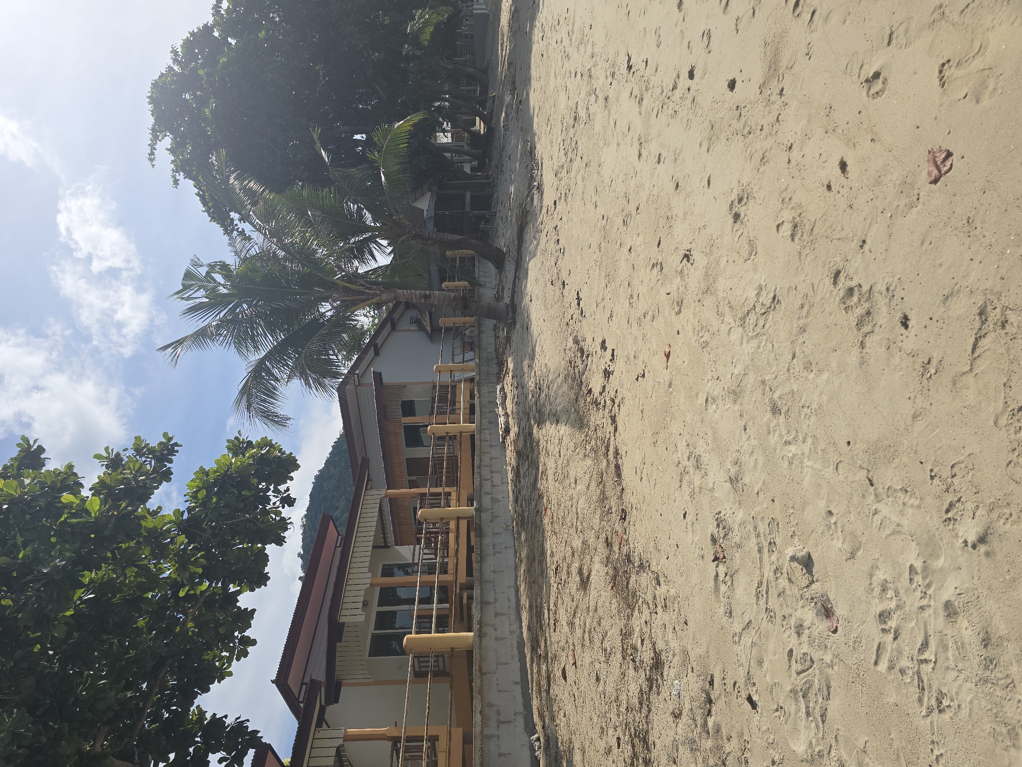 The image depicts a sunlit beach scene in Baan Lamai, Thailand. In the foreground, a sandy beach is visible, with numerous footprints and scattered leaves. Behind the beach, a low concrete wall supports a raised wooden deck or veranda that runs along the front of a building. This building appears to be a resort or guesthouse, with a light-colored facade, brown-tiled roof, and a railing along the veranda. Several palm trees and other lush green trees are scattered around the property and in the background. The sky is blue with scattered white clouds, and a lush green hillside is visible in the distance behind the buildings. The overall impression is one of a tropical beachside establishment.