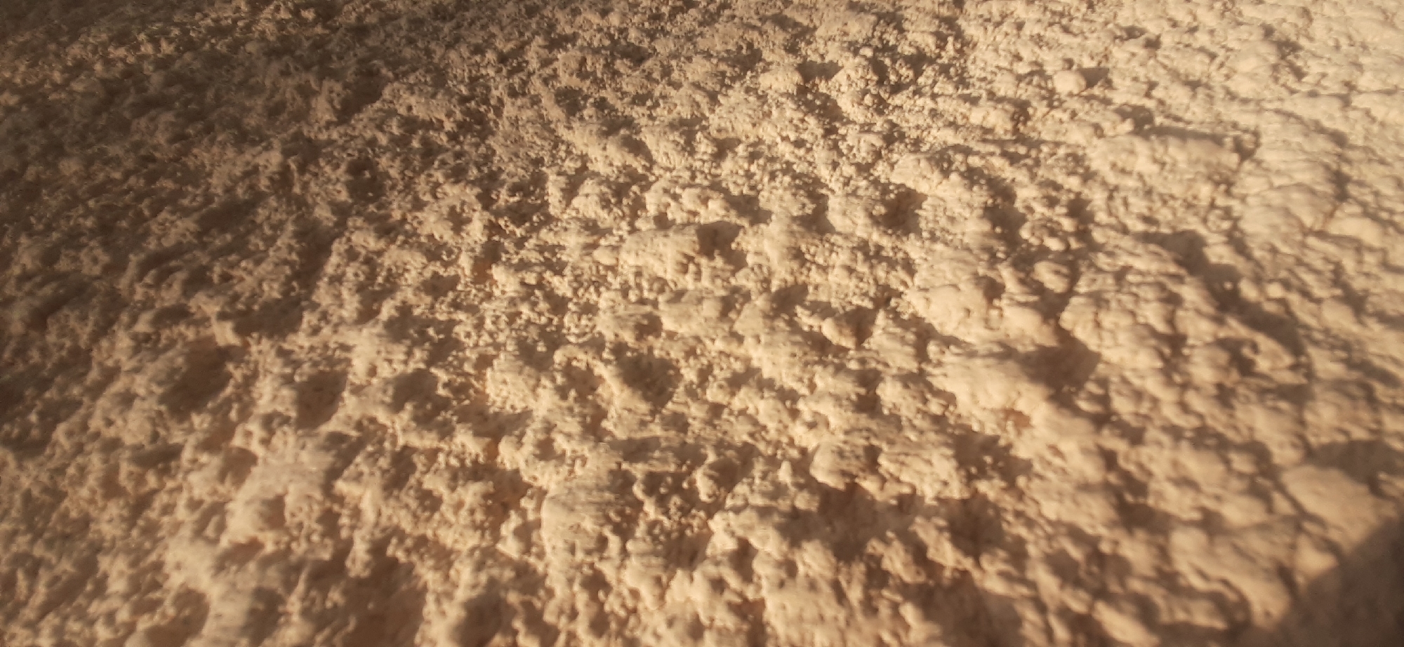 The image displays a close-up, angled view of a textured, light brown surface, possibly a wall or an object with a rough, granular finish. The lighting creates shadows that highlight the unevenness of the texture, with peaks and valleys of material visible. The color suggests it could be plaster, stucco, or a similar building material. The image is abstract and does not depict any specific location, people, or identifiable objects beyond the surface itself. There are no elements in the image that would allow for identification of its location in Yessentukskaya, Russia.