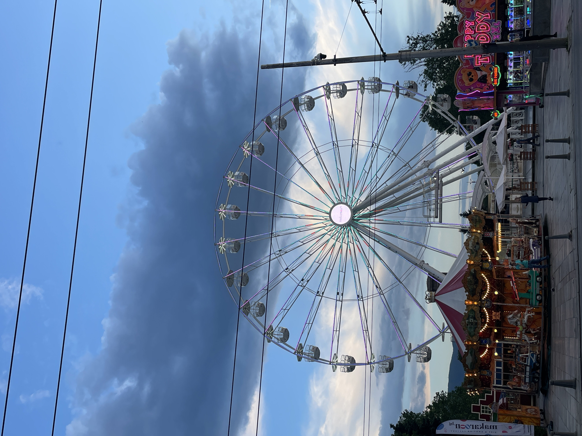 The image captures an evening scene at a lively amusement park or fairground in Sofia, Bulgaria. The sky is a mix of blues and grays, with a large, dark cloud dominating the left side, contrasting with lighter clouds and patches of blue sky on the right, indicating dusk or early evening.
The central subject is a large Ferris wheel, brightly illuminated with multicolored LED lights tracing its spokes and outlining its cabins. The center hub of the Ferris wheel displays text, which appears to be "makaroon." Below and to the right of the Ferris wheel is a classic carousel, also festively lit with warm, golden lights, featuring traditional horses and other ride figures.
Various fairground stalls and games are visible in the background on the right. One prominent, brightly lit stall features a colorful sign with cartoon characters and Cyrillic text that translates to "HAPPY KIDDY" and "BIG PRIZES," suggesting a game or children's attraction. Several people are scattered around the fairground, enjoying the evening, some standing near the rides and stalls.
In the foreground and extending across the sky, several black power lines or cables are visible, adding an urban or infrastructure element to the scene. At the bottom right, a vertical banner for "makaroon" is visible, with the additional text "DELIGHT YOUR DAY." The overall atmosphere is vibrant and celebratory, characteristic of an evening fair.