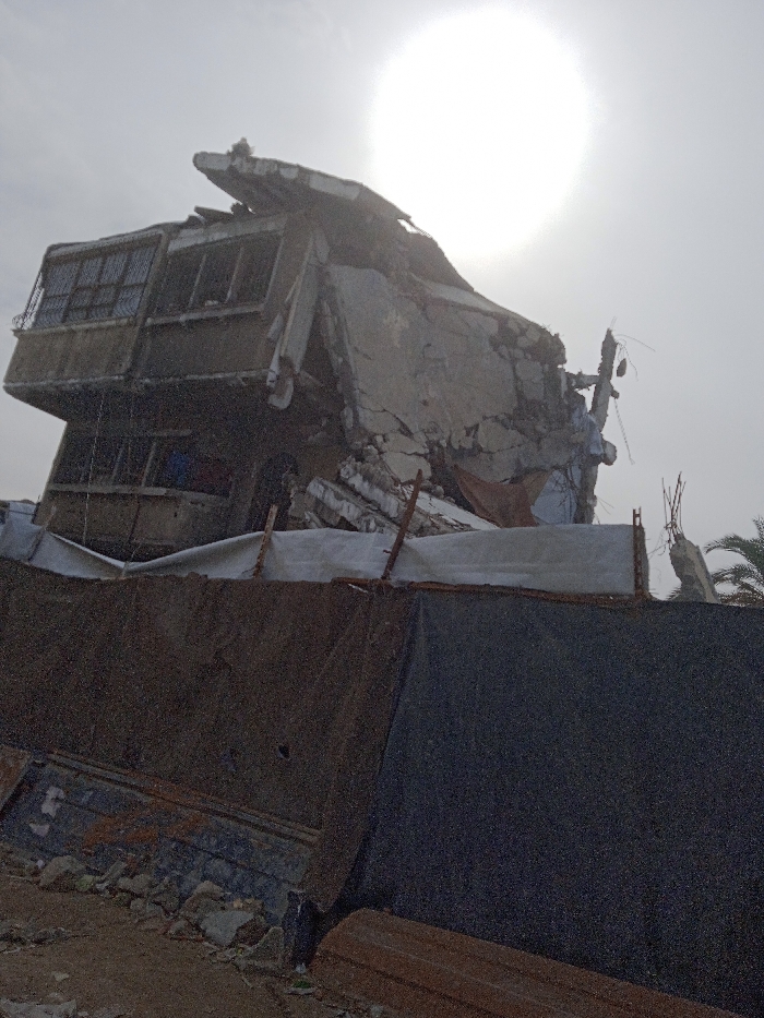 The image displays a severely damaged multi-story building located in Khan Yunis, Palestinian Territory, under a bright, hazy sky with the sun visible as a prominent, diffuse white disc in the upper right. The concrete structure exhibits extensive destruction, appearing partially collapsed and leaning. Its upper left section retains elements of a balcony with metal grates covering windows, while other windows on various levels are either open frames or contain broken glass. The main body of the building is characterized by fractured concrete slabs and exposed rebar, indicative of structural failure. An orange-brown fabric piece hangs from the debris.
In the foreground, a makeshift barrier is constructed from dark, corrugated metal sheets and a lighter-colored tarp, supported by visible stakes. The ground level in front of this barrier is composed of rough dirt and demolition debris, including chunks of concrete. A dark-colored cat is positioned near the base of the barrier on the left side. Several long, brown, rectangular objects, resembling wooden beams, lie on the ground in the lower right. Portions of a palm tree are discernible in the far right background. There are no human figures present, and no active interactions or discernible actions are occurring within the static scene.