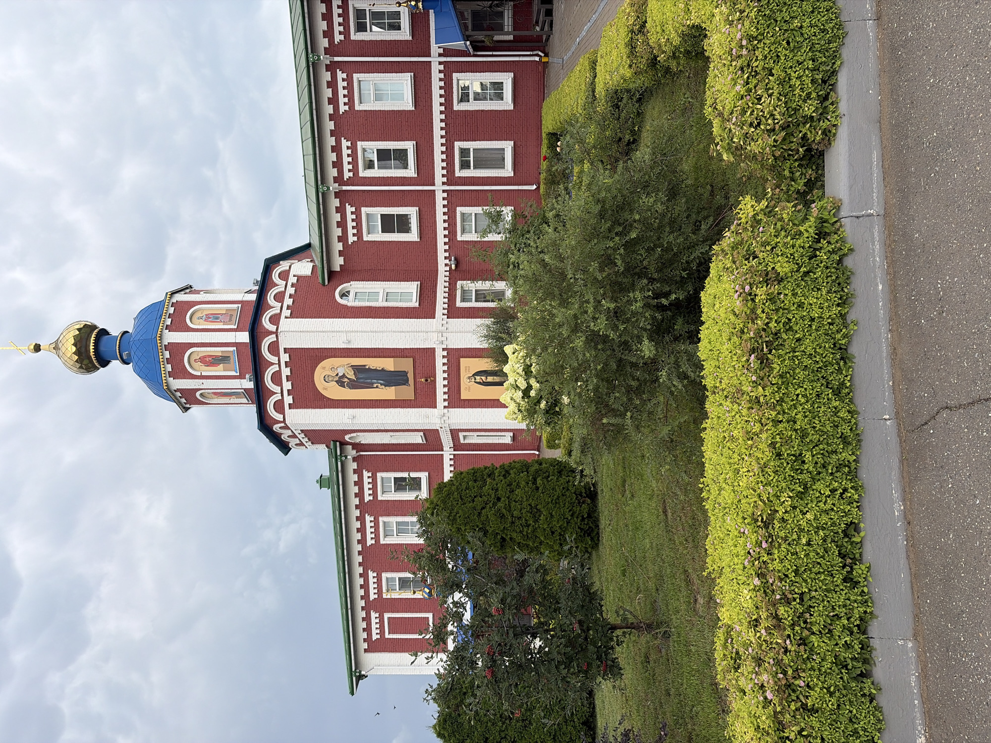 This is a medium shot, eye-level, exterior photograph of a red brick building with white trim, featuring a blue-domed tower topped with a golden cross. The building has multiple rectangular windows and ornate architectural details. On the tower, there are framed religious icons. In front of the building, there is a manicured garden with green bushes, a small tree with red berries, and flowering shrubs. The sky is overcast with clouds. The photograph appears to have been taken on a cloudy day. The building is identified as being located in the city of Norja, Russia.
