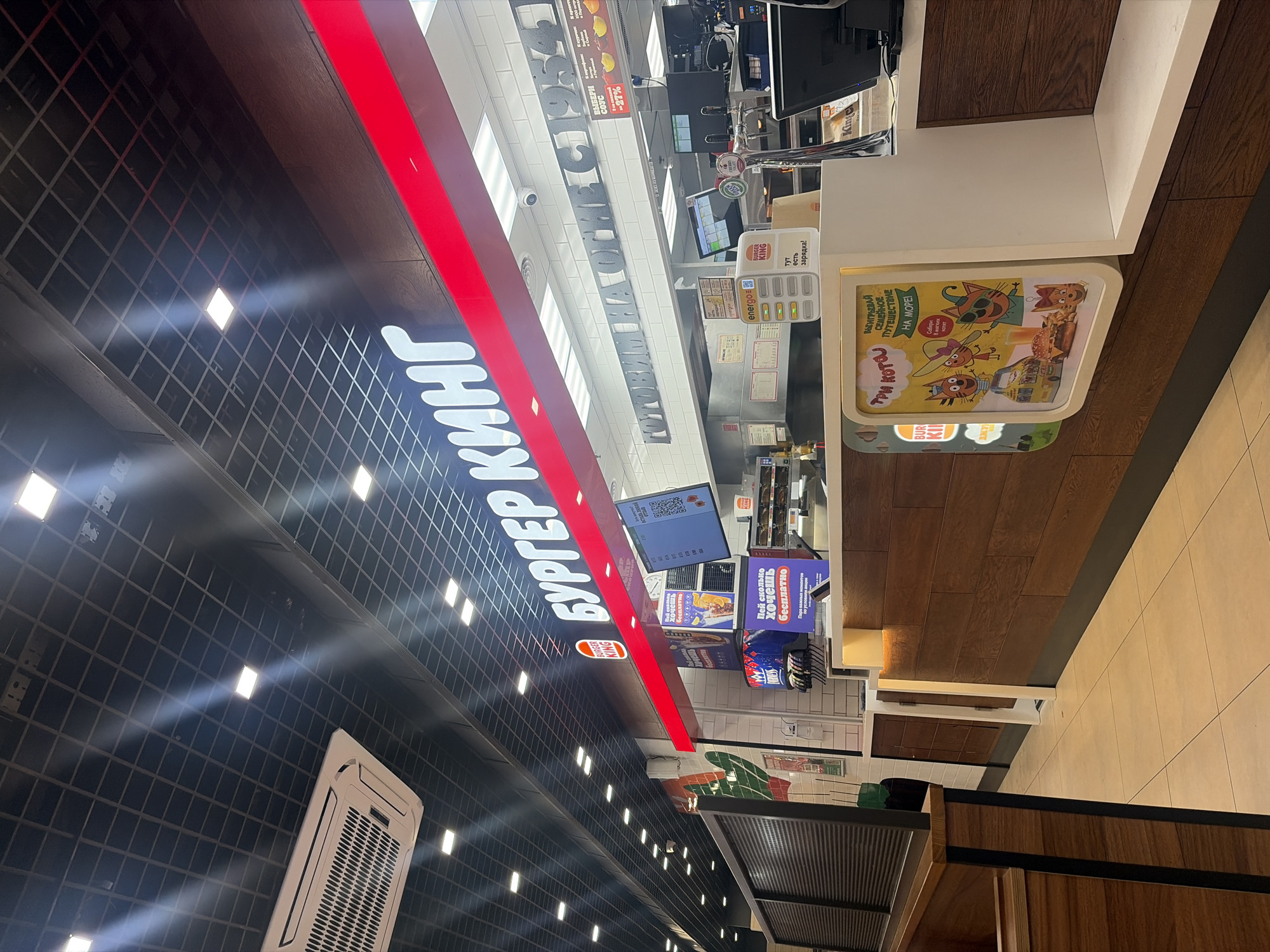 Here is a detailed description of the image:
**Environment:**
The photograph appears to be taken inside a Burger King restaurant, likely within a commercial establishment. The overall atmosphere is modern with a combination of industrial and warm design elements.
**Elements:**
* **Signage:** The Burger King logo and name, rendered in Cyrillic ("БУРГЕР КИНГ"), is prominently displayed with illuminated lettering. Other advertisements include posters featuring cartoon cats for a promotion in Russian.
* **Interior:** The interior design includes a ceiling with a black grid-like pattern with integrated lighting. Wood panels and modern light fixtures contribute to the aesthetics.
* **Ordering Area:** The image captures part of the counter and ordering area, displaying menu screens, point-of-sale equipment, and beverage dispensers.
* **Objects:** A charging station for mobile phones labeled "energo" and "BURGER KING" is visible.
* **People:** There are no clearly discernible individuals visible in the photograph.
**Location:**
Given the Cyrillic signage (Бургер Кинг), the language of the promotional materials, and the general architectural style, it's likely this Burger King restaurant is located in a Russian-speaking area, potentially in the city of Saint Petersburg, Russia.