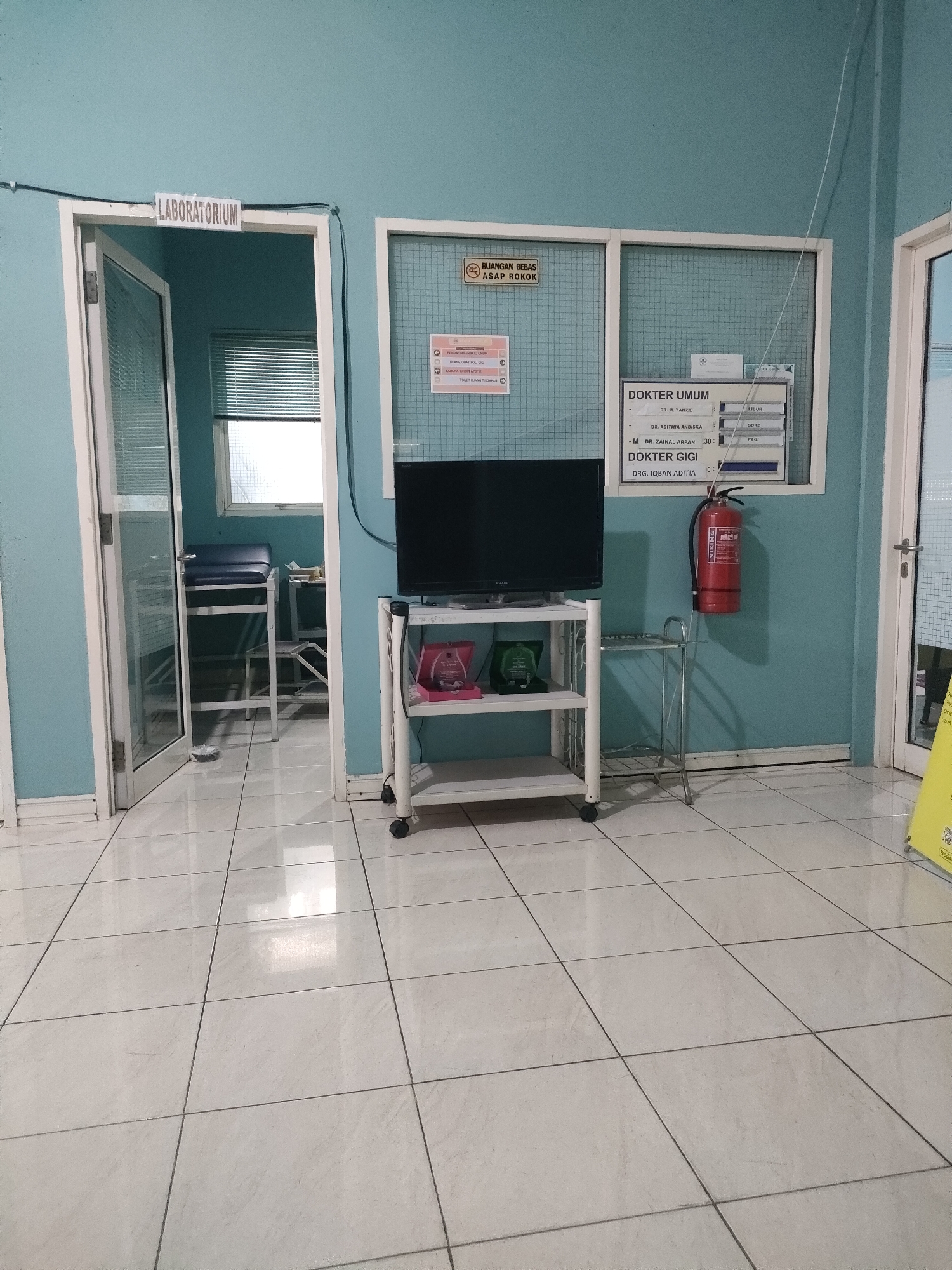 The image depicts the interior of a medical facility, likely a clinic or hospital, in Sungailiat, Indonesia. The walls are painted a pale blue, and the floor is tiled with white square tiles. A door on the left, labeled "LABORATORIUM," opens into a room with a window and blinds. In the center of the image, a white shelf unit with wheels holds a black television, two pink boxes, and a green award. Adjacent to the shelf is a silver metal stand. On the wall above the shelf, a schedule board lists doctors' names and their working hours. To the right, a red fire extinguisher is mounted on the wall. Further to the right, another door, partially visible, leads to a different area. The overall environment appears clean and organized, suggesting a functional medical space.