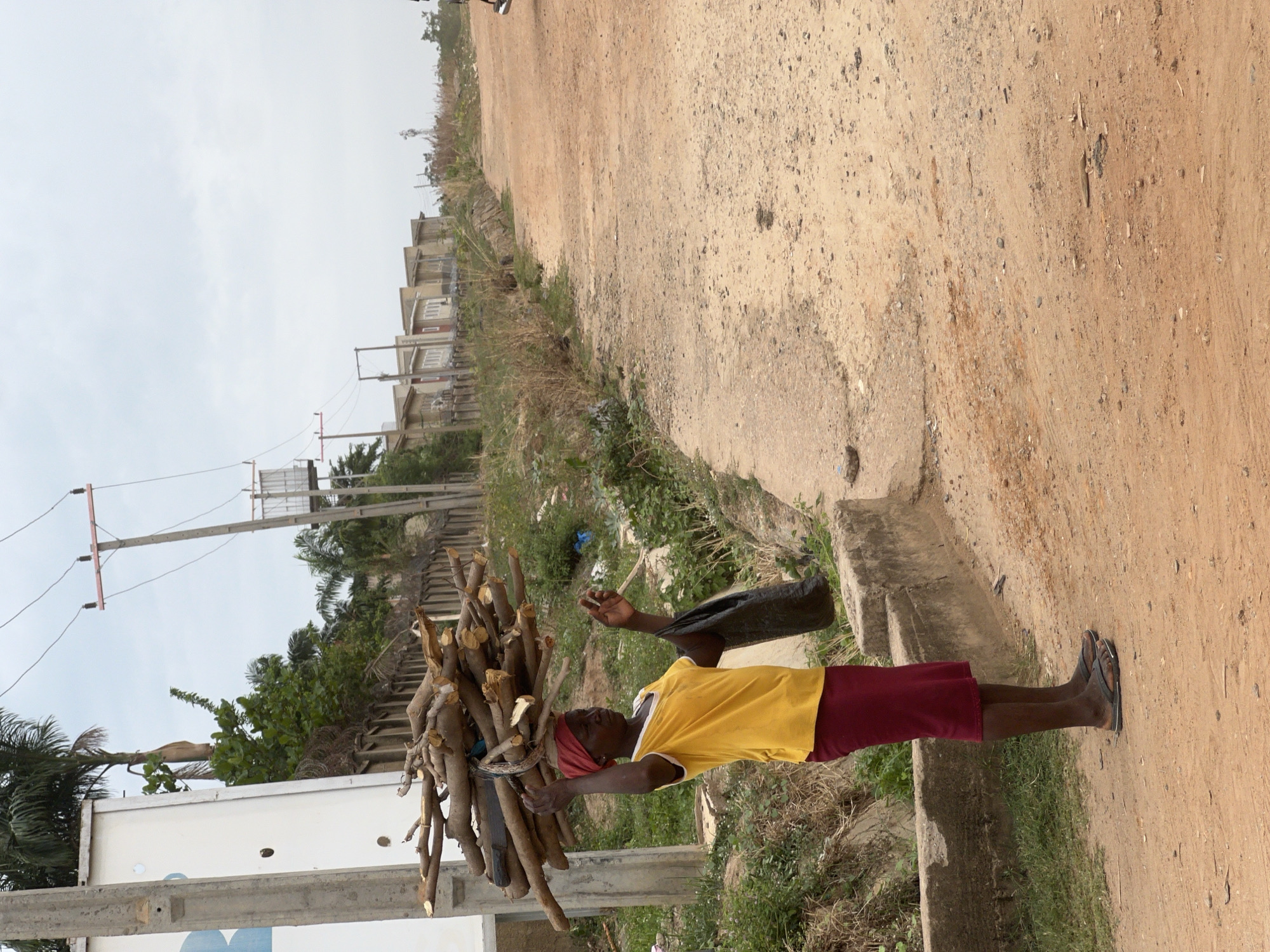 A woman is carrying a bundle of firewood on her head along a dirt road. She is wearing a yellow t-shirt, a red skirt, and sandals. She also has a red head covering. The background shows buildings, trees, and power lines. The location appears to be a developing area with unpaved roads, possibly on the outskirts of Abuja, Nigeria.