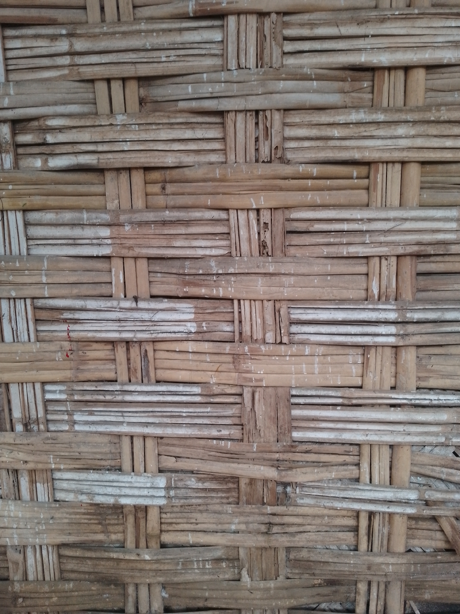 This is a close-up, eye-level, full-frame photograph of a woven bamboo wall in Kauman, Indonesia. The bamboo is a light brown and tan color, with some sections appearing whitewashed. The weaving pattern is a simple over-under technique, creating a textured surface. The bamboo pieces are of varying thickness and some show signs of wear and tear, with cracks and peeling. There are small white speckles and some reddish-brown marks visible on the surface of the bamboo, likely from paint or other debris. The lighting is even, indicating an overcast day or shaded area, with no harsh shadows. The focus is sharp across the entire image, highlighting the intricate details of the woven material. No people or other objects are visible in the frame, making the bamboo wall the sole subject.