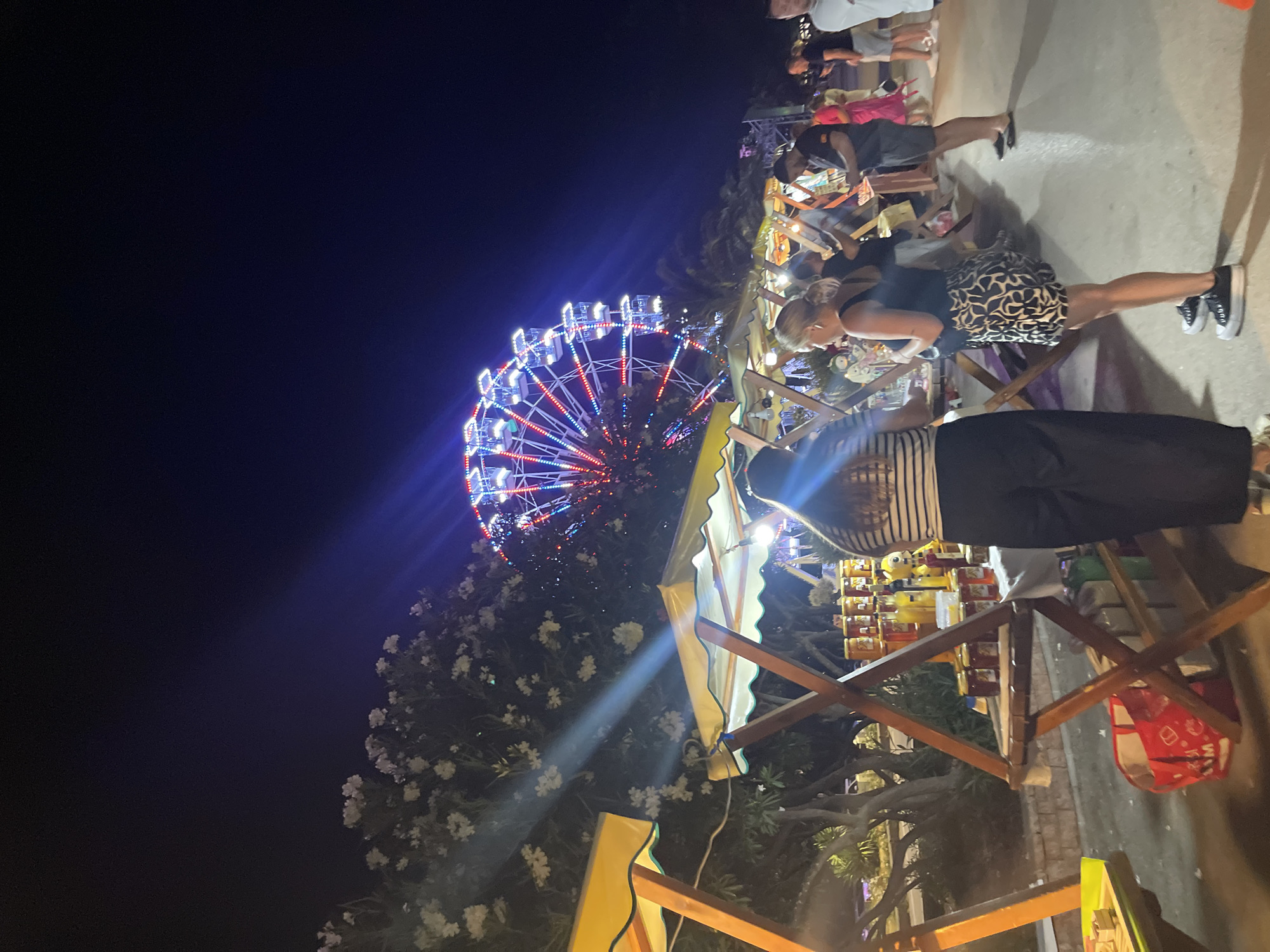 A night scene at an outdoor market in Bar, Montenegro, features a brightly lit Ferris wheel in the background. In the foreground, several market stalls with yellow awnings display various goods, including jars of honey. People are browsing at the stalls. To the left, a person wearing a striped shirt and dark pants stands near a stall. To the right, a woman in a black tank top and patterned skirt is looking at a stall, accompanied by a man in a dark shirt and shorts. Other individuals are visible further back, with a white building and trees also present in the environment. The ground appears to be paved.