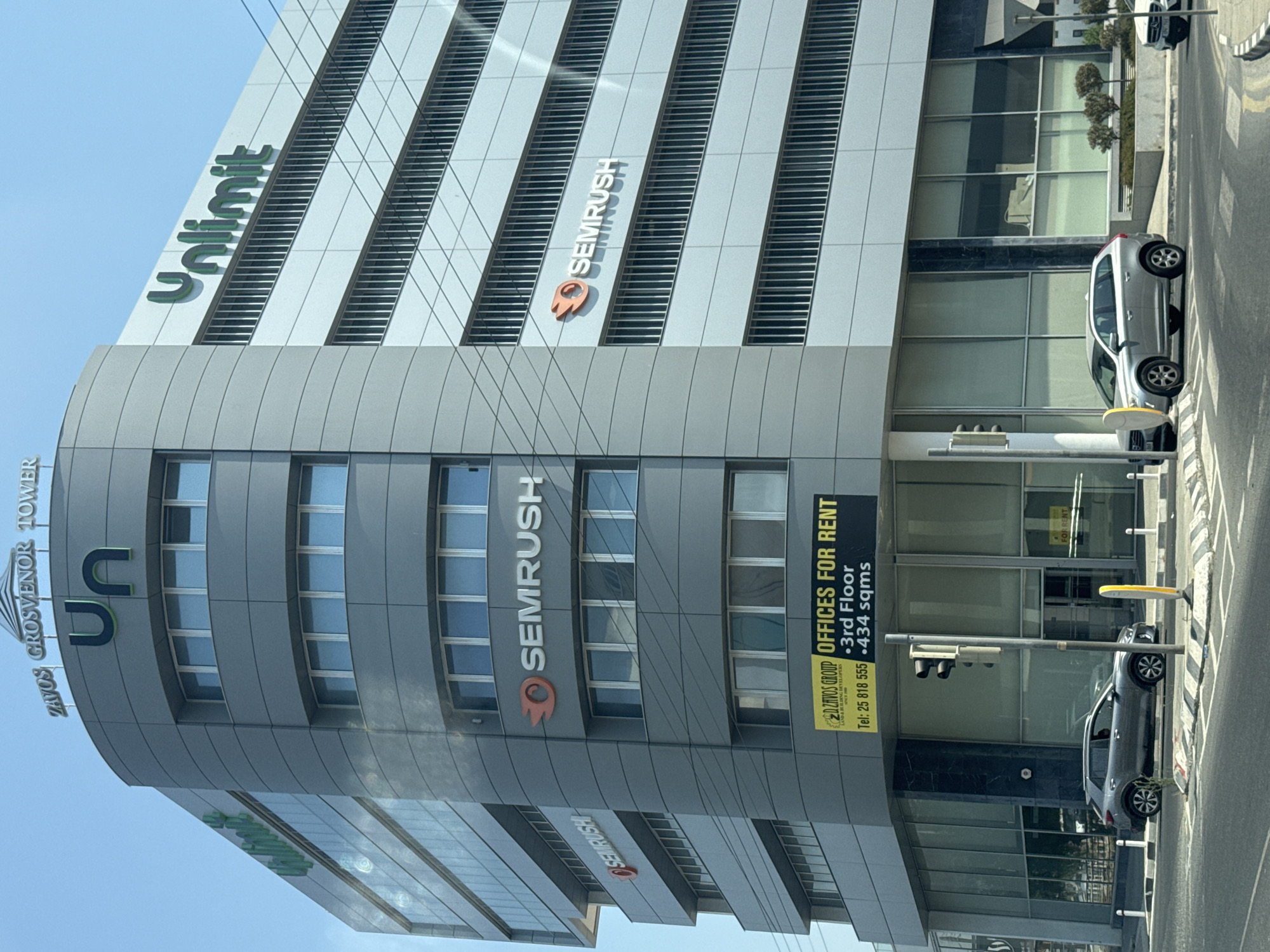 A modern, multi-story office building located on a street in Limassol, Cyprus is depicted. The building is primarily grey with prominent signage. On the top of the building, the words "ZAVOS GROSVENOR TOWER" are visible. The building features several logos: "Un" in a large green font on the curved section of the building, and "Unlimit" in green on the right side. Multiple instances of the "SEMRUSH" logo, a stylized orange flame and text, are present on different floors.
A yellow sign on the lower part of the building advertises "OFFICES FOR RENT" with contact details and floor information. The ground floor has large glass windows, and a traffic light is visible. Two cars are parked on the street in front of the building: a grey SUV on the left and a silver SUV on the right. The environment appears to be a commercial district with a clear sky.