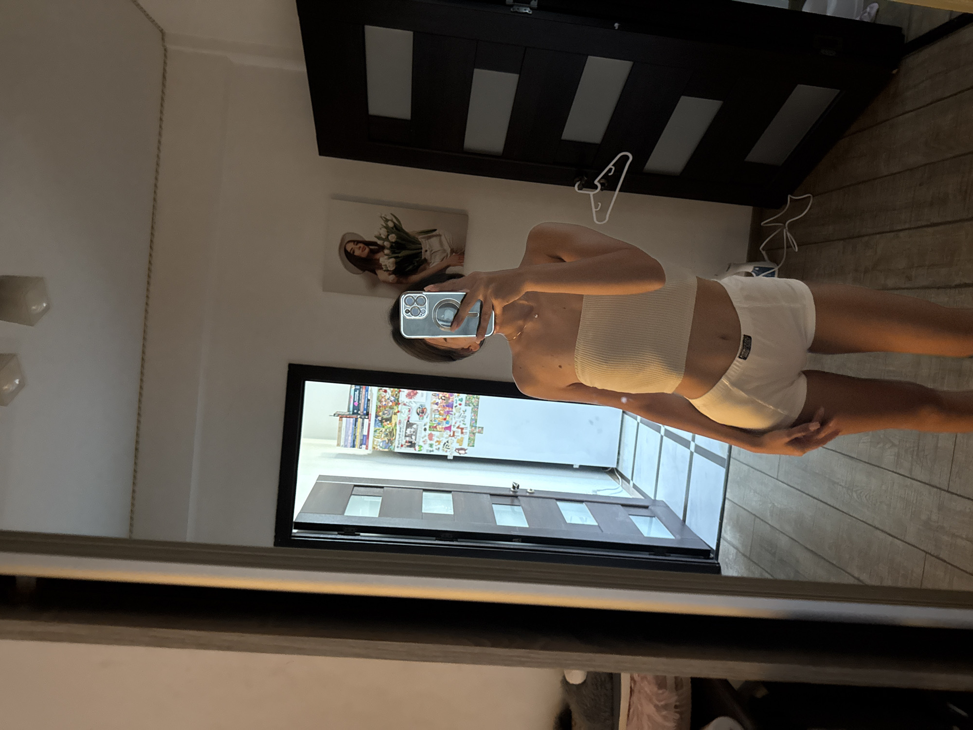 A young woman is standing in a room, taking a mirror selfie. She is wearing a white bandeau top and white boxer-brief style shorts. The room has wooden flooring and a large mirror. To the left of the mirror, a doorway leads to another part of the apartment, revealing a hallway with tiled flooring and a door with glass panes. On the wall behind the woman, a framed picture of a woman wearing a hat and holding flowers is visible. To the right, a dark door with horizontal glass panels stands closed. A white clothes hanger is hooked onto the frame of this door. The overall impression is of a personal space, possibly a bedroom or living area within an apartment in Chișinău, Moldova.