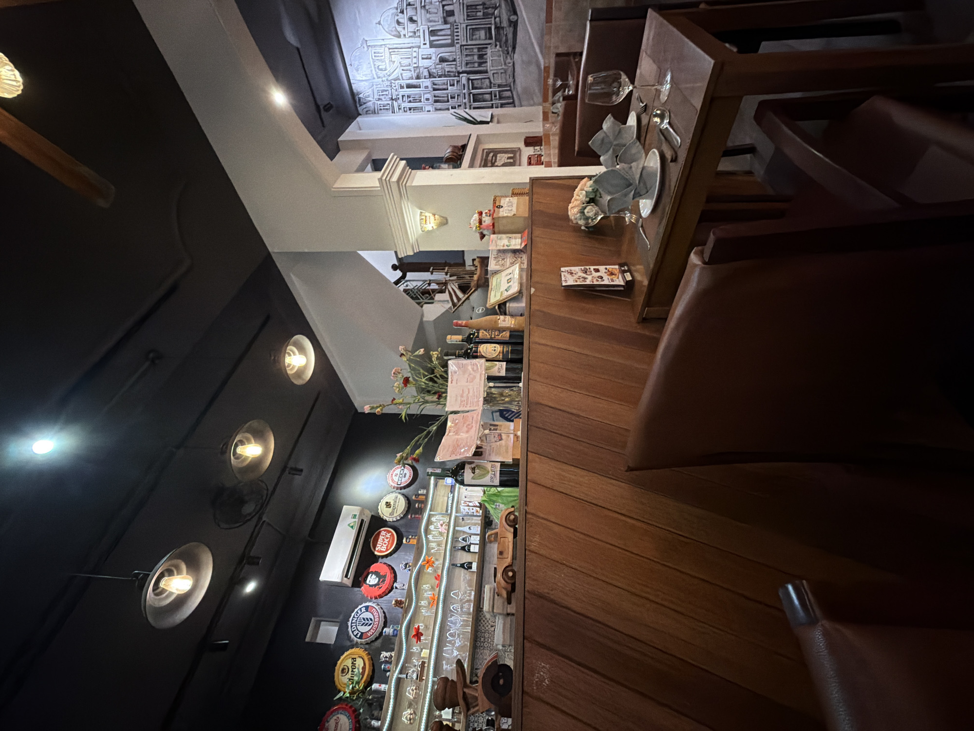 The photograph depicts the interior of a restaurant in Thủ Đức, Vietnam. The scene is dimly lit, with warm light emanating from pendant lamps on a black ceiling. The restaurant features wooden paneling on the walls and a bar area showcasing various beer caps and bottles. On the bar, there are decorative wooden car models and a collection of glassware. A shelf behind the bar is stocked with bottles and other decorative items.
In the foreground, a table is set for a meal, with white plates, folded napkins, cutlery, and wine glasses. Two brown leather chairs are positioned at the table. In the background, a mural of a European cityscape adorns the wall, adding a touch of artistry to the establishment. A staircase leads to an upper level, and a large window provides a glimpse of the exterior. The overall ambiance suggests a cozy and intimate dining experience.