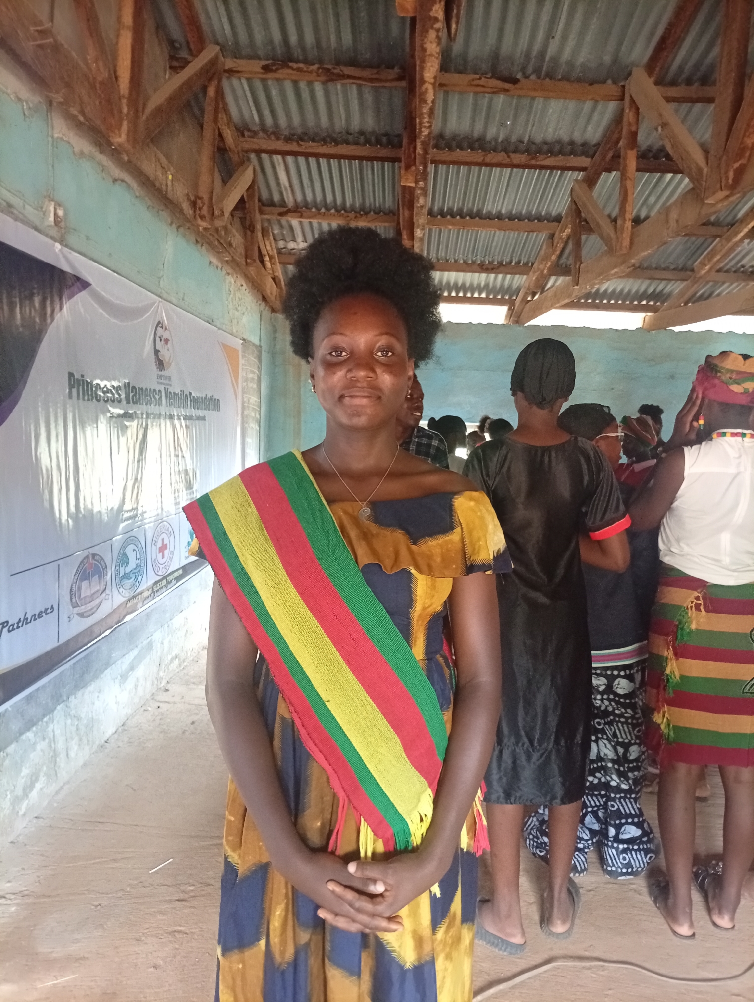 The image captures a group of people, primarily women, standing indoors in what appears to be a community hall or event space. The primary subject in the foreground is a young woman with dark, curly hair, wearing a multi-colored off-the-shoulder dress with a yellow, navy, and brown pattern. She also wears a sash with horizontal stripes of red, yellow, green, and black. Her hands are clasped in front of her, and she is looking directly at the camera with a neutral expression.
Behind her, several other individuals are visible. To her right, a person in a black outfit with red trim is facing away from the camera. Further to the right, a woman is partially visible, wearing a white top and a colorful striped skirt. More people are standing in the background, some partially obscured.
The environment is characterized by a rustic, unfinished ceiling made of corrugated metal supported by wooden beams. The walls are painted a light blue. A large banner is visible on the left wall, featuring text and logos, including "Princess Vanessa Yemilo Foundation" and "Nigerian Red Cross." The floor is dirt or compacted earth. The overall scene suggests an event or gathering in a rural or semi-rural setting. The location is described as Unknown, Unknown.