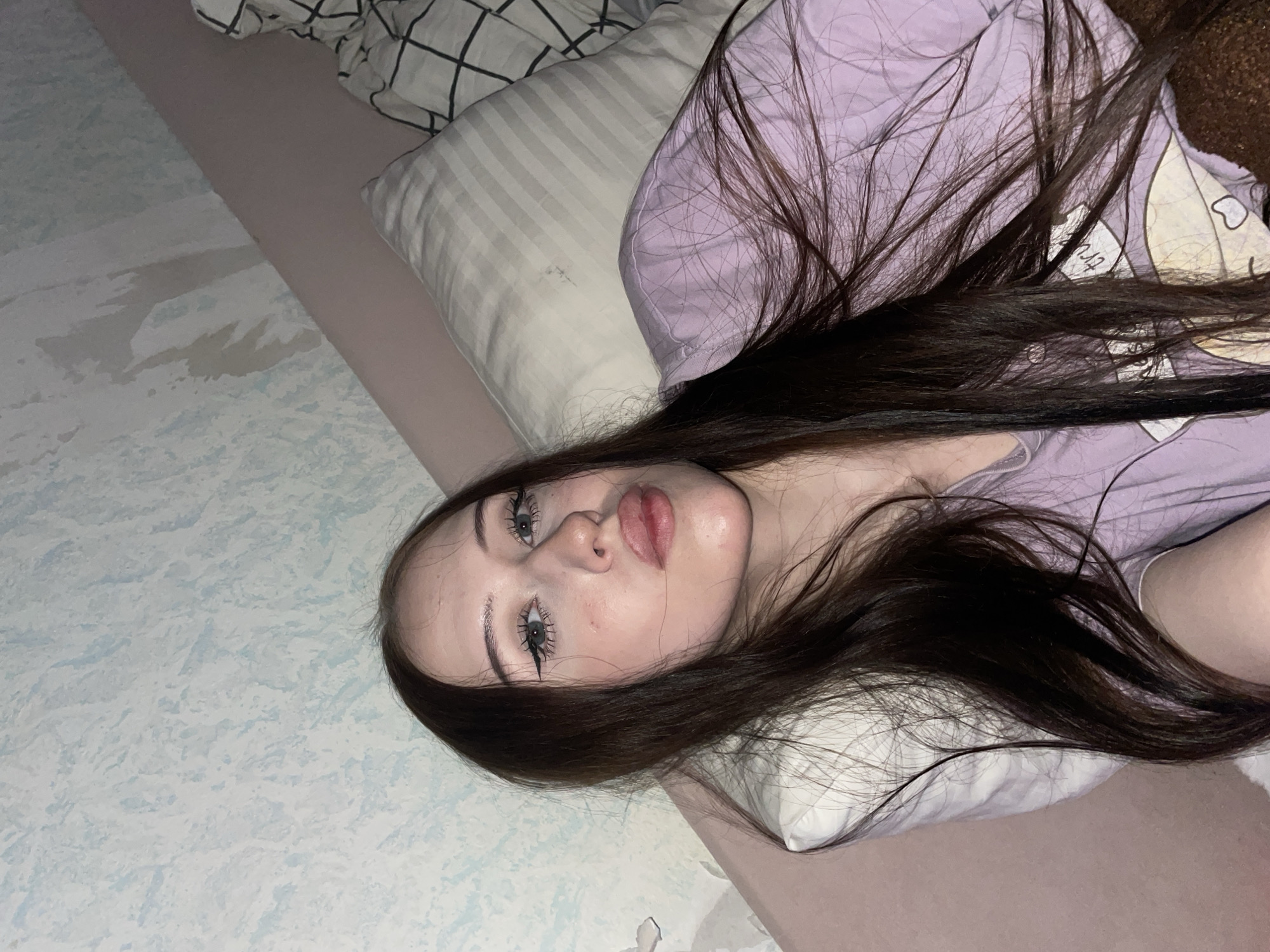 The media file is a close-up, portrait-oriented photograph of a young woman lying on a bed. She has long, dark brown hair spread across her shoulders and over a light purple garment, possibly pajamas. The woman is looking directly at the camera, with her eyes open and her lips slightly parted. Her makeup includes dark eyeliner and mascara, and her skin appears smooth.
The setting is indoors, on a bed with various pillows and bedding. To the left of the frame, a light blue and white patterned sheet or comforter is visible. There are two pillows behind the woman: one with thin grey and white stripes, and another with a checkered pattern of black and white lines. The overall lighting suggests it might be nighttime or indoors with artificial light, creating a soft glow on her face. No specific weather conditions or external location cues are visible. The woman's expression could be interpreted as relaxed or perhaps a bit sleepy, given the context of lying in bed. There is no visible text in the image. The location context of Votkinsk, Russia, is provided but not visually evident within the image itself.