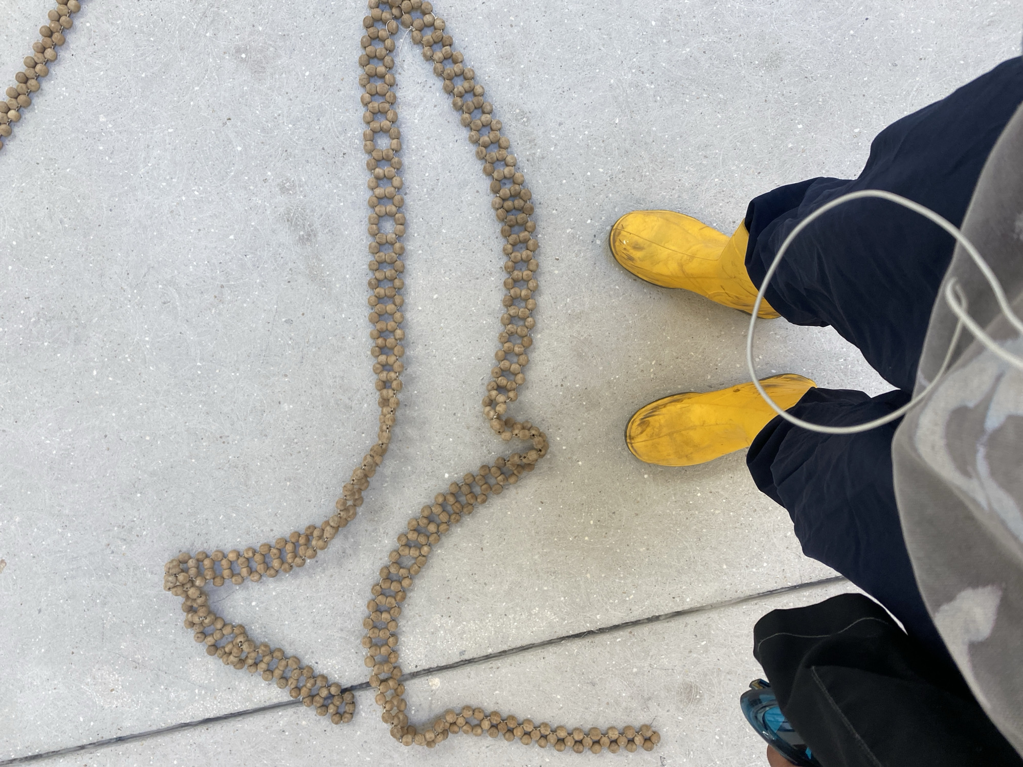 The image captures an overhead view of an outdoor urban scene, likely a sidewalk or paved area in Paris, France. The primary subjects are a person's lower body and a long, winding decorative chain made of small beads, laid out on the light grey ground.
The person is wearing dark blue or black trousers and striking, bright yellow, possibly rubber, boots. One foot is firmly on the ground, while the other leg is slightly bent, suggesting a standing or momentarily paused posture. A white cable, likely from wired headphones or earphones, loops around the person's torso. In the bottom right corner, a portion of what appears to be another person's dark clothing and a blue shoe or bag is visible, though less clearly.
The most prominent object is the elongated chain, intricately crafted from numerous small, light brown, spherical beads. These beads are strung together in a double-strand pattern, creating a flat, flexible ribbon-like structure. The chain forms a meandering, abstract shape across the concrete surface, spanning from the top left of the frame down towards the bottom center. Its texture contrasts with the smooth, speckled pavement.
The time of day appears to be daytime, with bright, even lighting suggesting a clear sky. There are no discernible emotions or explicit activities taking place beyond the implied observation of the bead chain. No visible text is present in the image.
