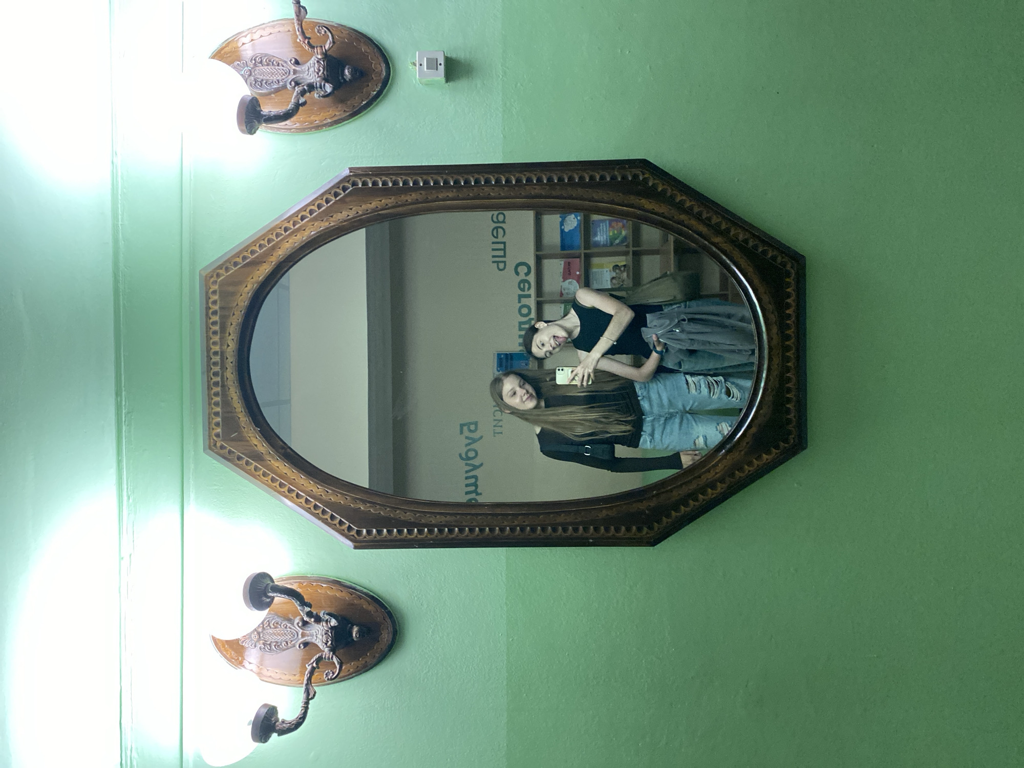 The image shows a medium shot of two young women reflected in an ornate, octagonal mirror. The mirror is mounted on a light green wall that transitions to a slightly darker green to the right. Two vintage-style wall sconces with wooden bases and metal accents are visible, one above and to the left of the mirror, and another below and to the left.
In the reflection, both women are smiling and appear to be taking a selfie with a phone. The woman on the left has long blonde hair and is wearing a black tank top and ripped blue jeans. The woman on the right has long brown hair, also wearing a black top and blue jeans. Behind them, the reflection reveals a bookshelf filled with books and some decorative items.
Visible text in the reflection includes what appears to be Greek lettering on a box or frame, reading "ΕΛΛΗΝΙΚ" and "ΓΕΜΠ". There is also text on the spine of a book in the background, partially obscured. The lighting suggests it is daytime, possibly indoors in a well-lit room, as indicated by the bright wall sconces. The overall mood is casual and cheerful. The location context provided is Votkinsk, Russia.