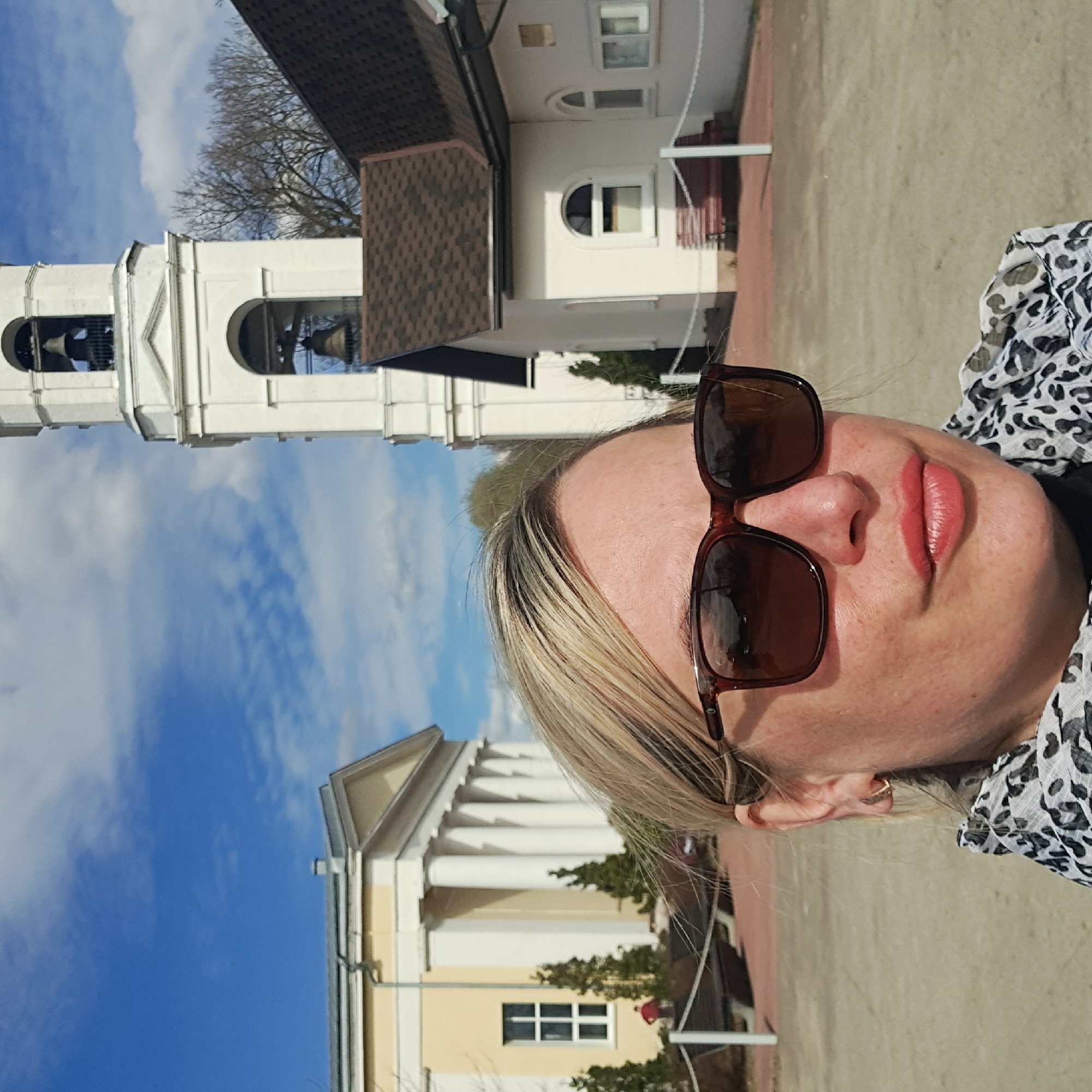 A selfie photograph shows a blonde woman wearing sunglasses. Behind her are two buildings against a blue sky with white clouds. The building on the left is a bell tower with a white facade and arched openings revealing bells. The building on the right has a light yellow facade, white columns, and a triangular pediment. The setting is Ivashevo, Russia.