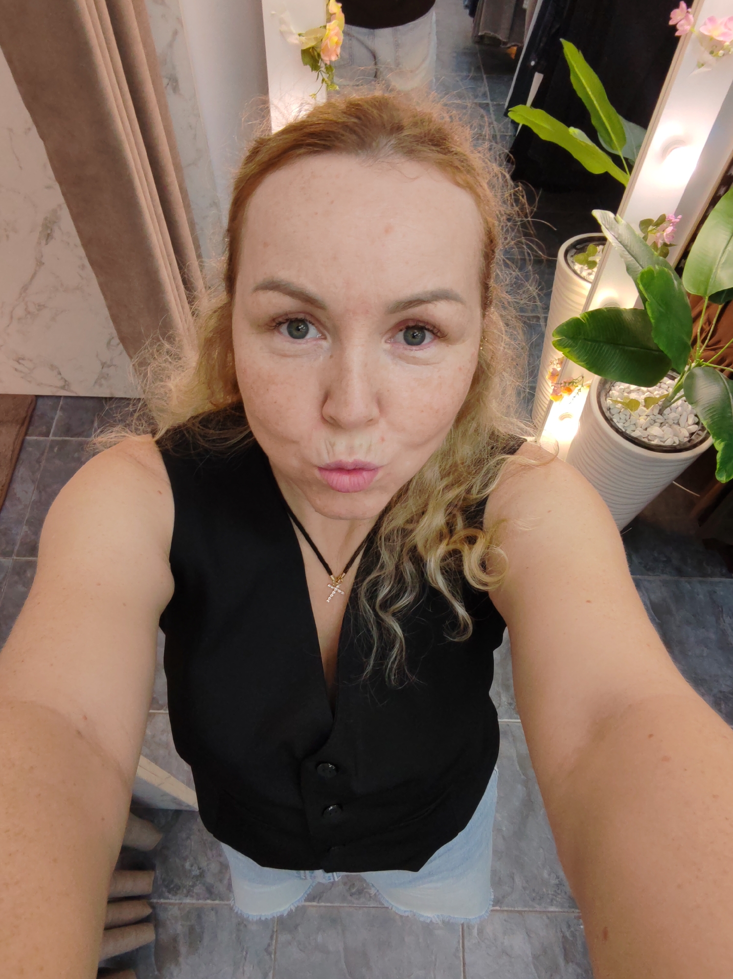 A woman with blonde, wavy hair and freckles is taking a selfie. She is wearing a black vest over a light blue denim skirt and a necklace with a cross pendant. Her lips are pursed, and she is looking at the camera. The background shows a tiled floor, a curtain, a plant in a pot, and what appears to be a lighted mirror. The location is the city of Chaikovsky, Russia, though the specific setting within the city is not identifiable.