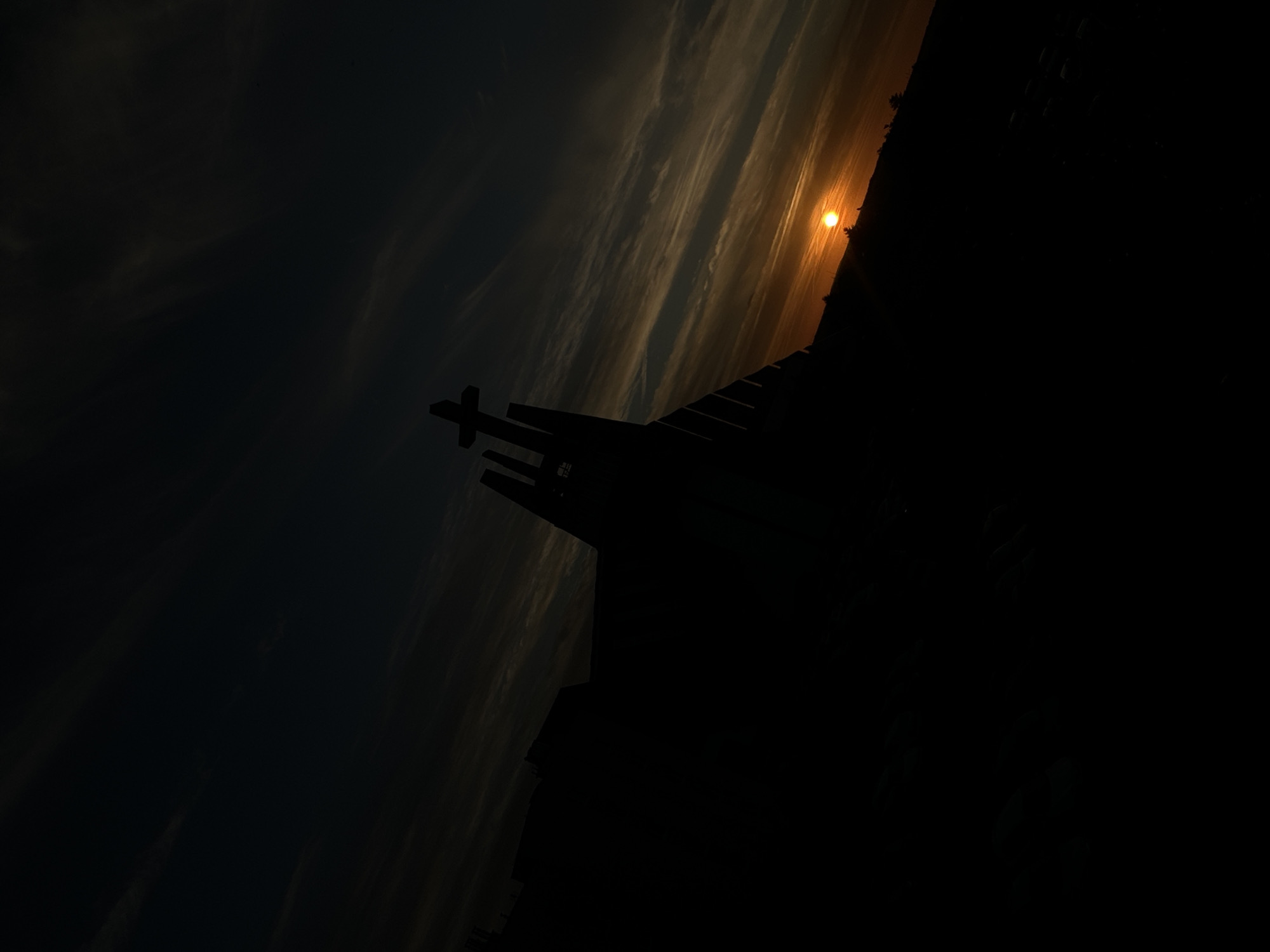 The image is a silhouetted view of the Krzyż Milenijny (Millennium Cross) and a portion of the surrounding landscape at sunset. The cross, a prominent landmark in Świdnica, Poland, is silhouetted against a sky streaked with orange and dark clouds. The sun is a bright orange orb low on the horizon, casting a warm glow. The dark silhouette of the cross and the foreground suggest it is a significant structure, likely a monument or a religious building, positioned on a hill. The tilt of the image enhances the dramatic effect of the sunset.